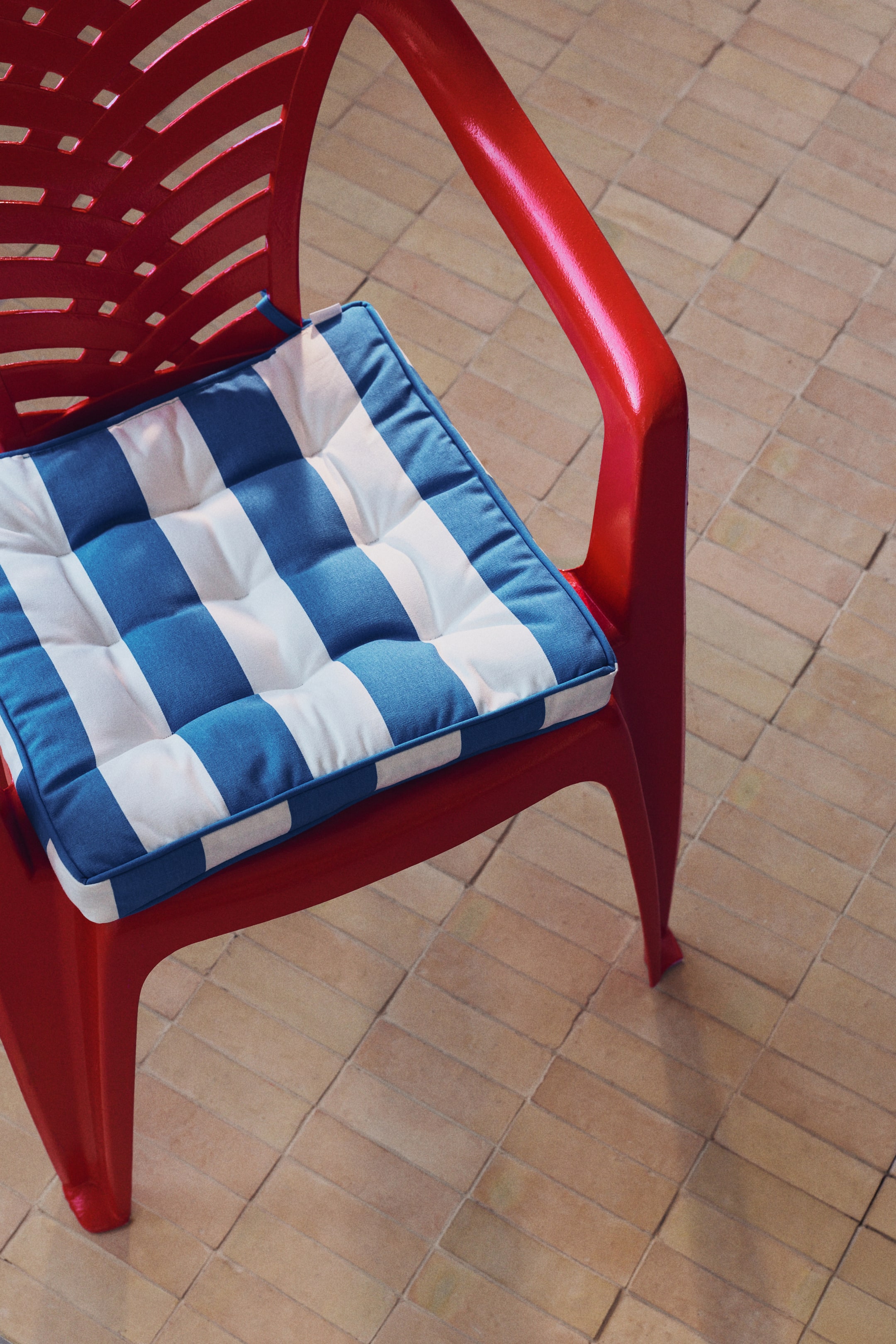 Ampliar la imagen: Una silla de plástico rojo brillante, vista desde un ángulo alto, presenta un respaldo con listones y reposabrazos curvados. Un cojín cuadrado con rayas azules y blancas, que muestra capitoné y ribeteado a lo largo de sus bordes, descansa sobre el asiento.