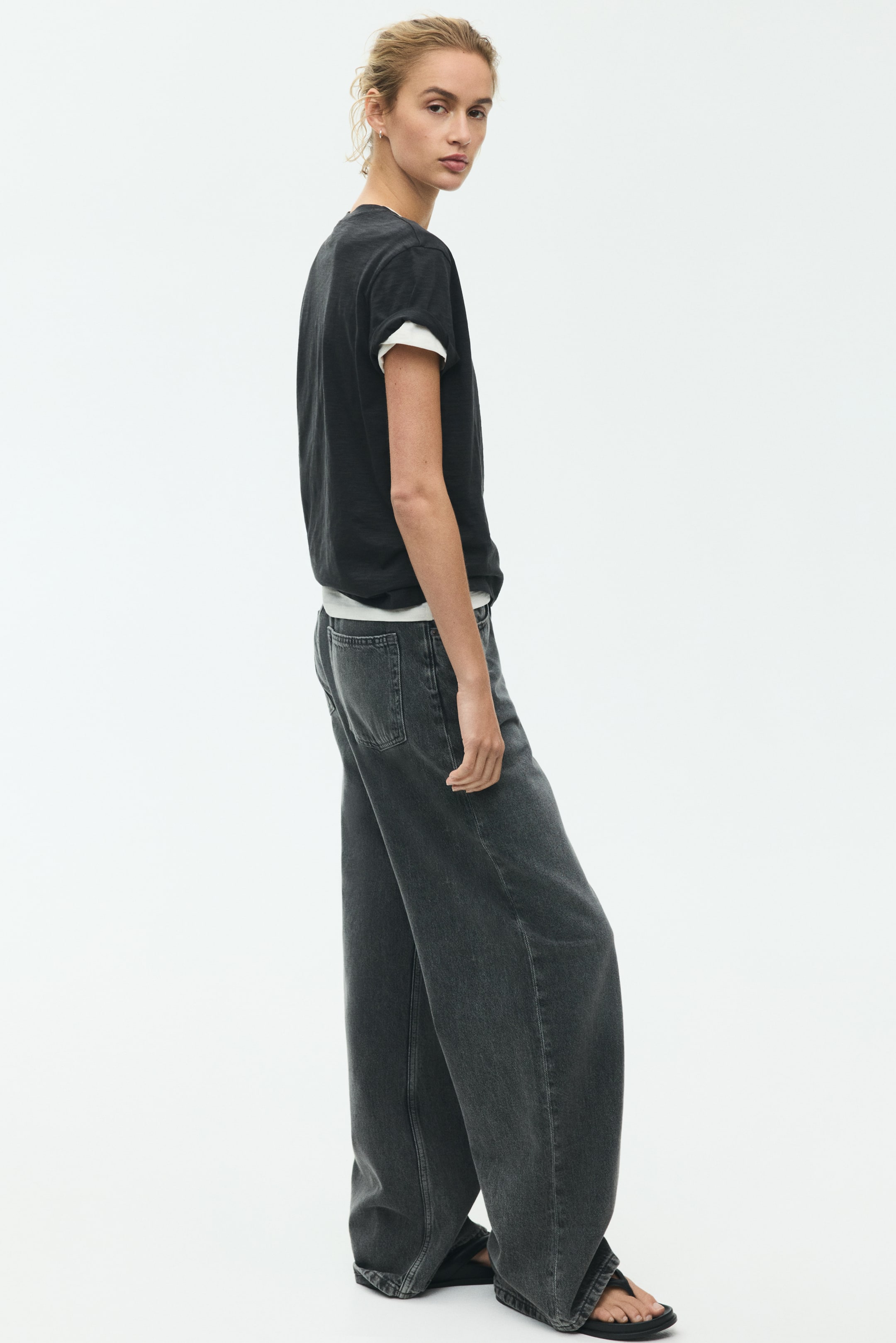 View larger image: A charcoal gray short-sleeve t-shirt with a relaxed fit is showcased from a slight side angle, revealing a white layer at the neckline and rolled sleeve cuffs. Charcoal gray wide-leg jeans are also displayed, highlighting visible back pockets.