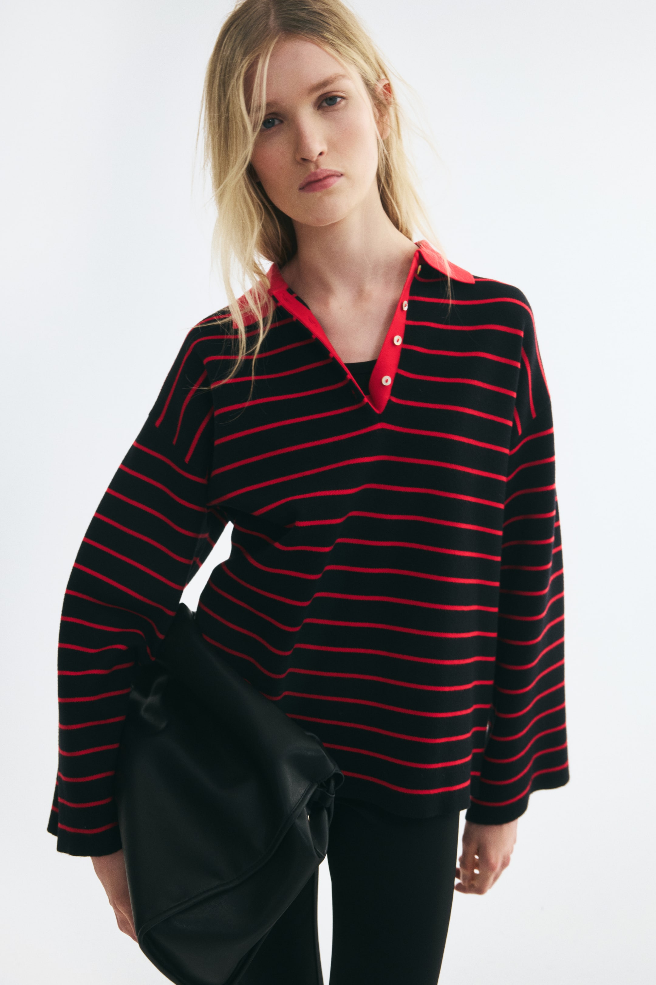 View larger image: An oversized polo shirt is draped loosely in a front-facing view, featuring horizontal black and red stripes. The shirt displays dropped shoulders, wide long sleeves, and a contrasting solid red collar with an open, partially buttoned placket.