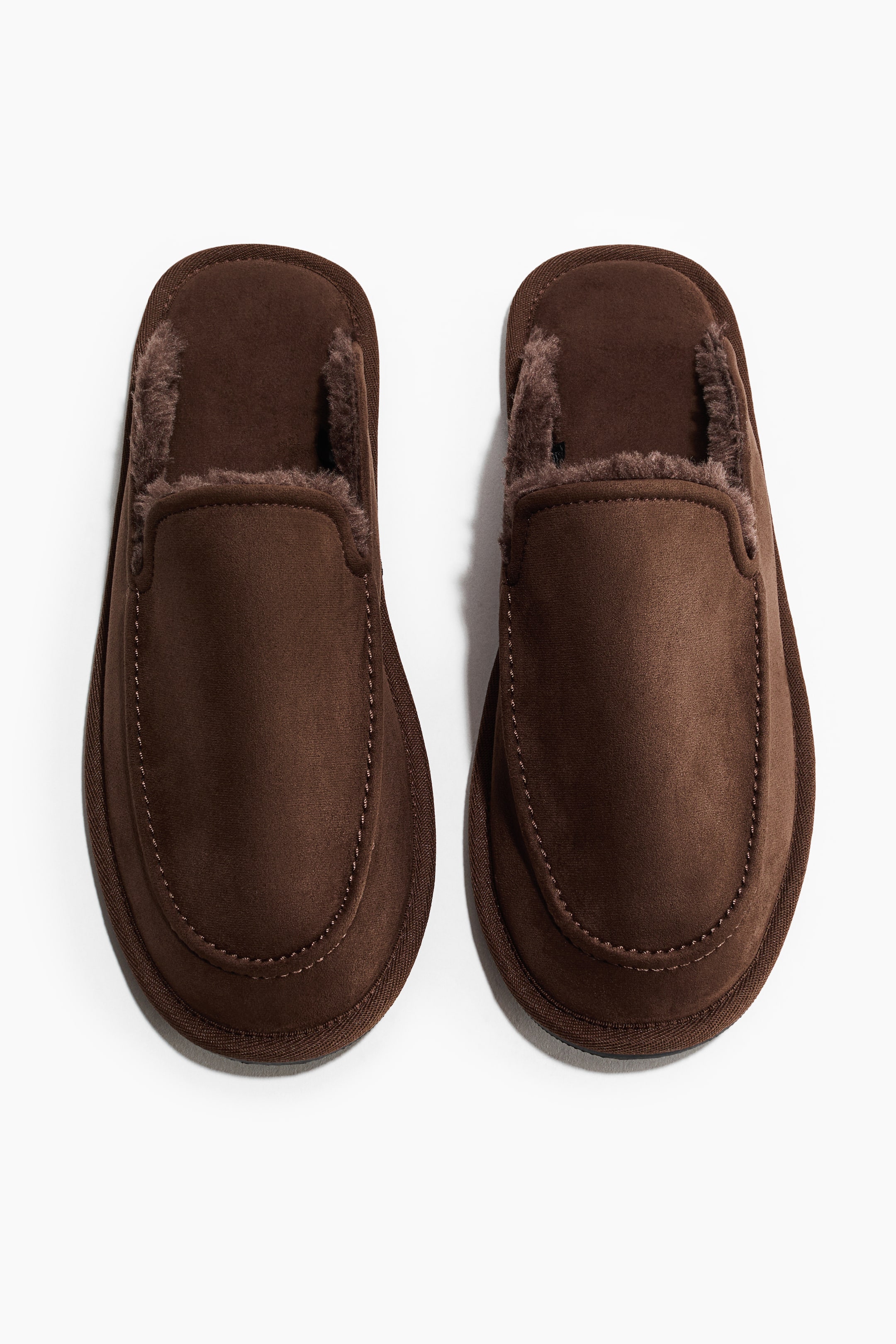 View larger image: A pair of brown slip-on slippers is facing forward, seen from above, each featuring a moccasin-style toe with visible stitching and a shearling-like lining visible around the opening.