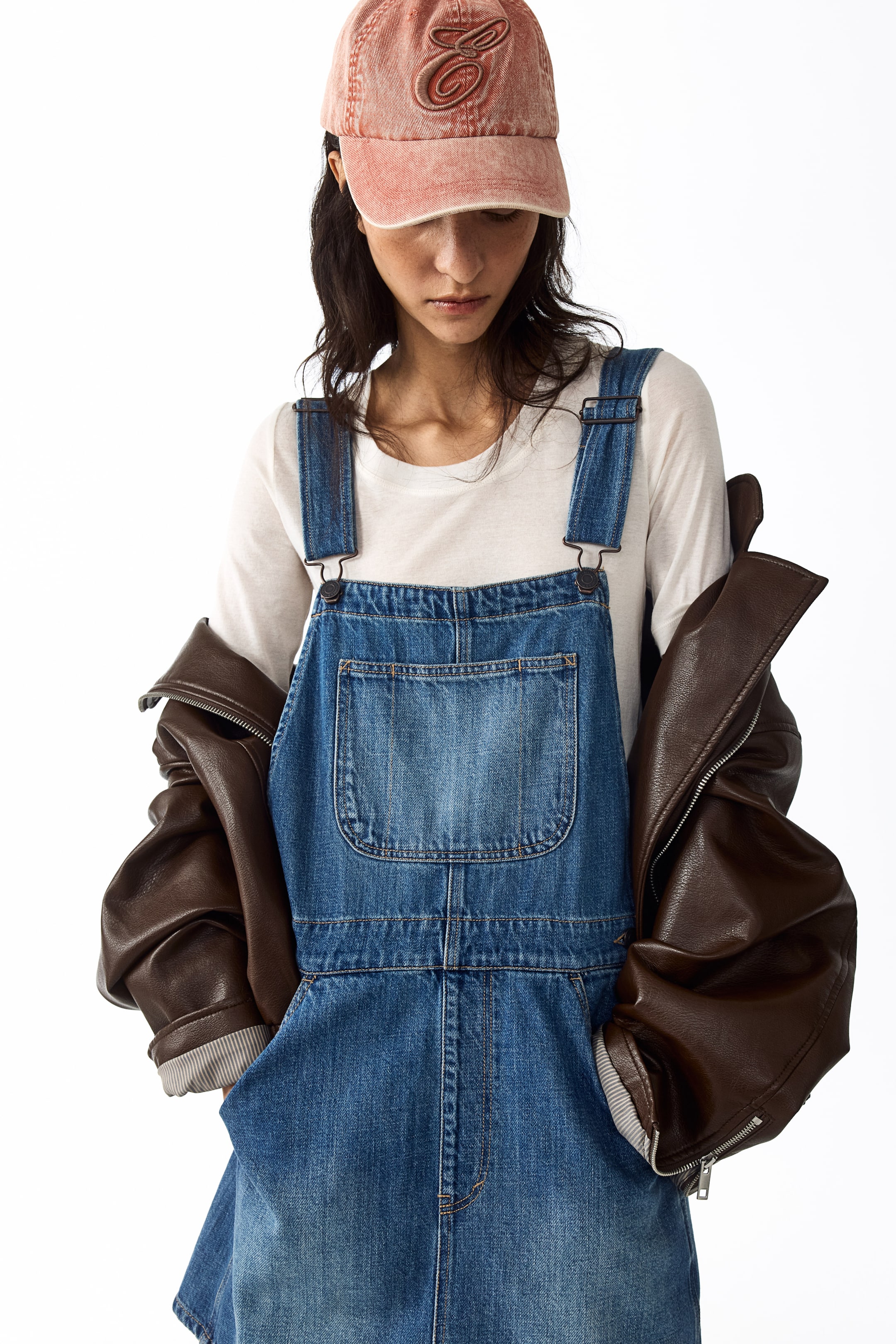Voir une image plus grande: Une robe salopette en denim bleu présente un devant à bavette avec une grande poche plaquée rectangulaire et des bretelles ajustables. Elle est portée sur un t-shirt blanc à manches longues.