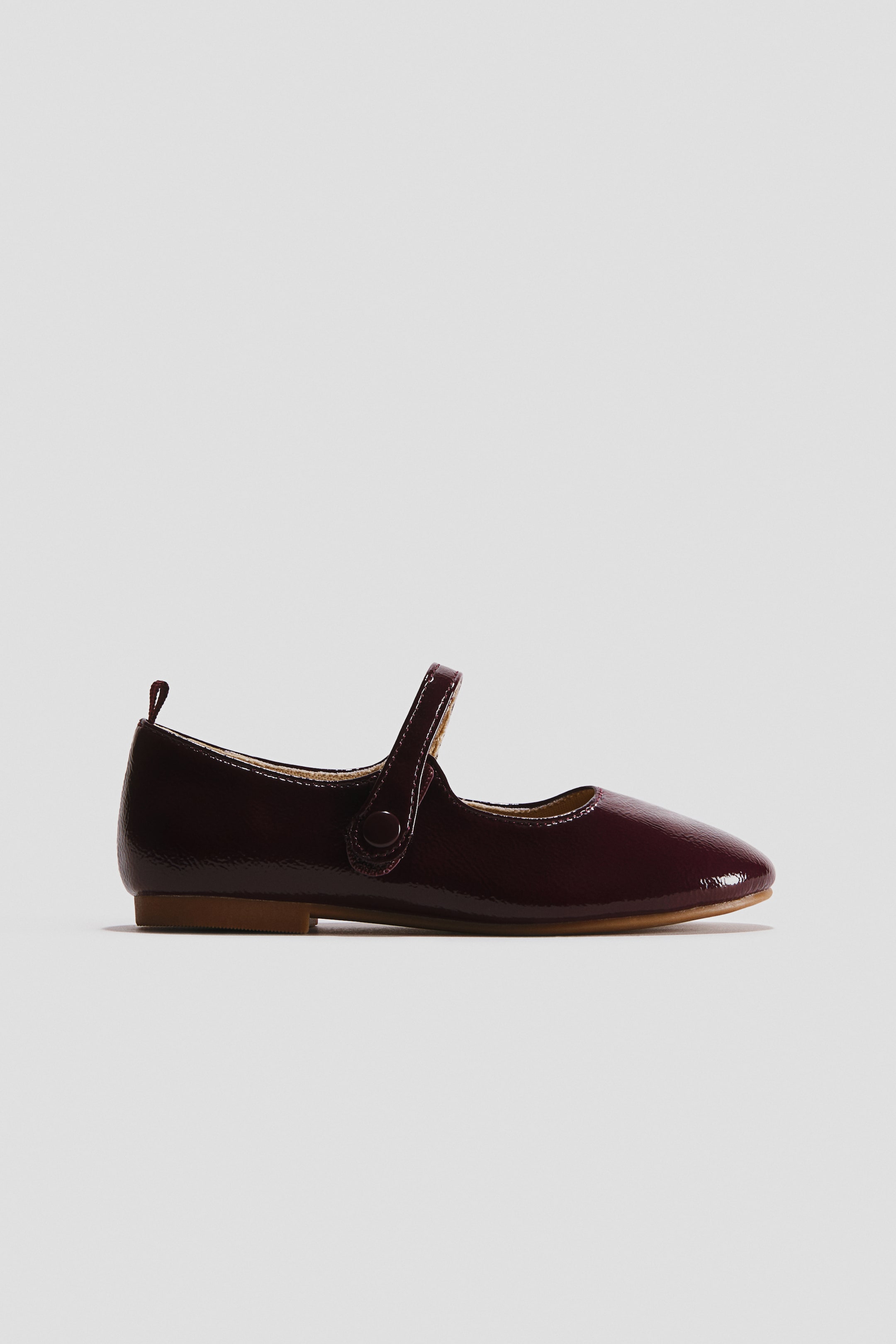 View larger image: A burgundy Mary Jane shoe is shown in side profile facing right, featuring a glossy patent finish and an instep strap with a decorative button cover. The shoe is designed with a small pull loop at the back of the heel and a flat, light-brown sole.