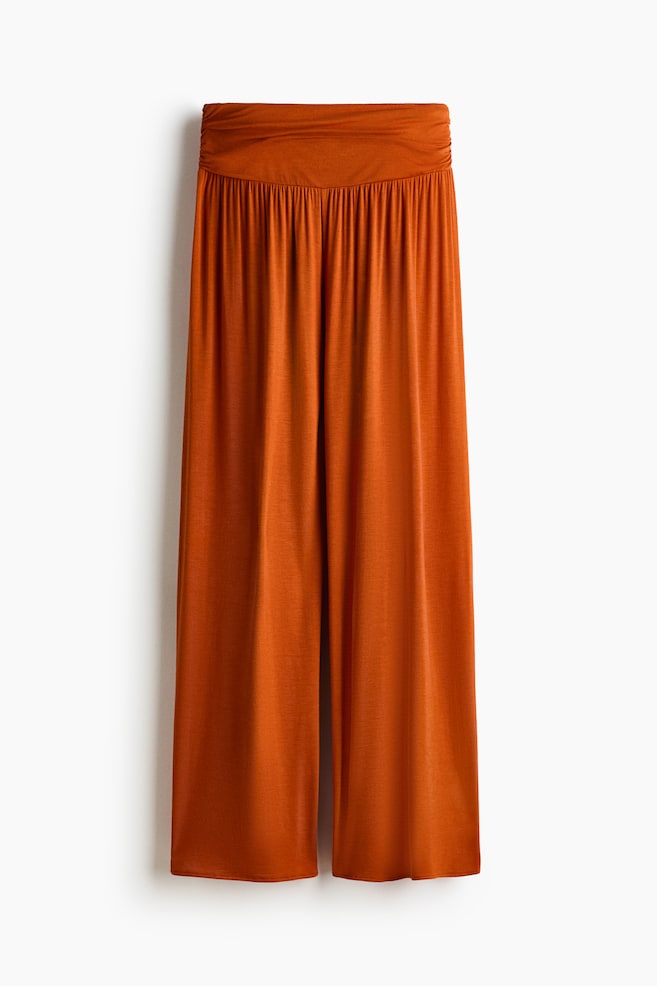 A pair of burnt orange pants is displaying its front, featuring a wide, smooth waistband with gathering directly beneath it, and flowing wide legs. - 1