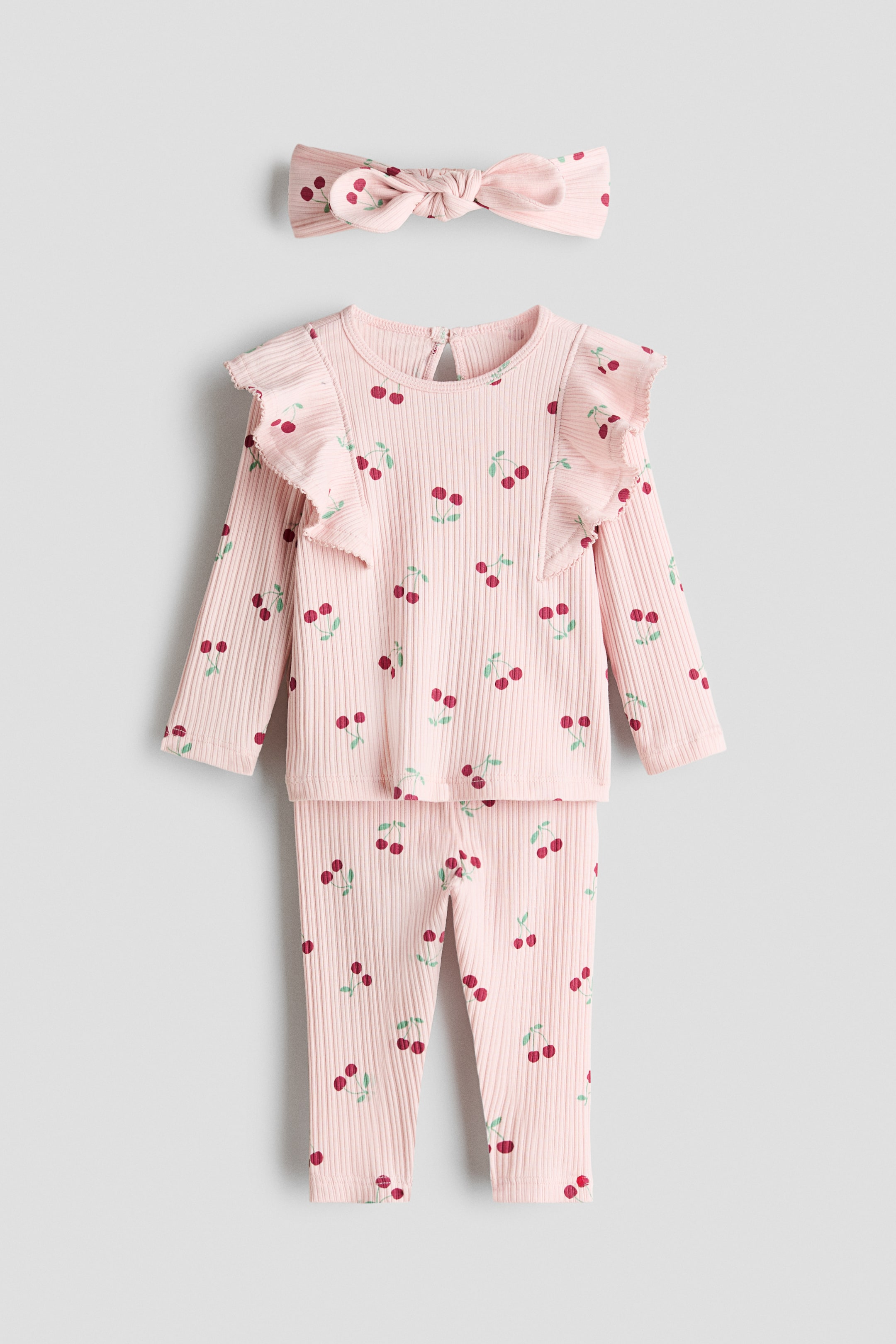 View larger image: A pink ribbed knit three-piece baby set is displayed, showing the FRONT. The set includes a long-sleeve top with ruffled shoulders and a round neckline with a keyhole opening at the back, matching full-length pants, and a headband tied in a bow, all featuring an all-over cherry print.