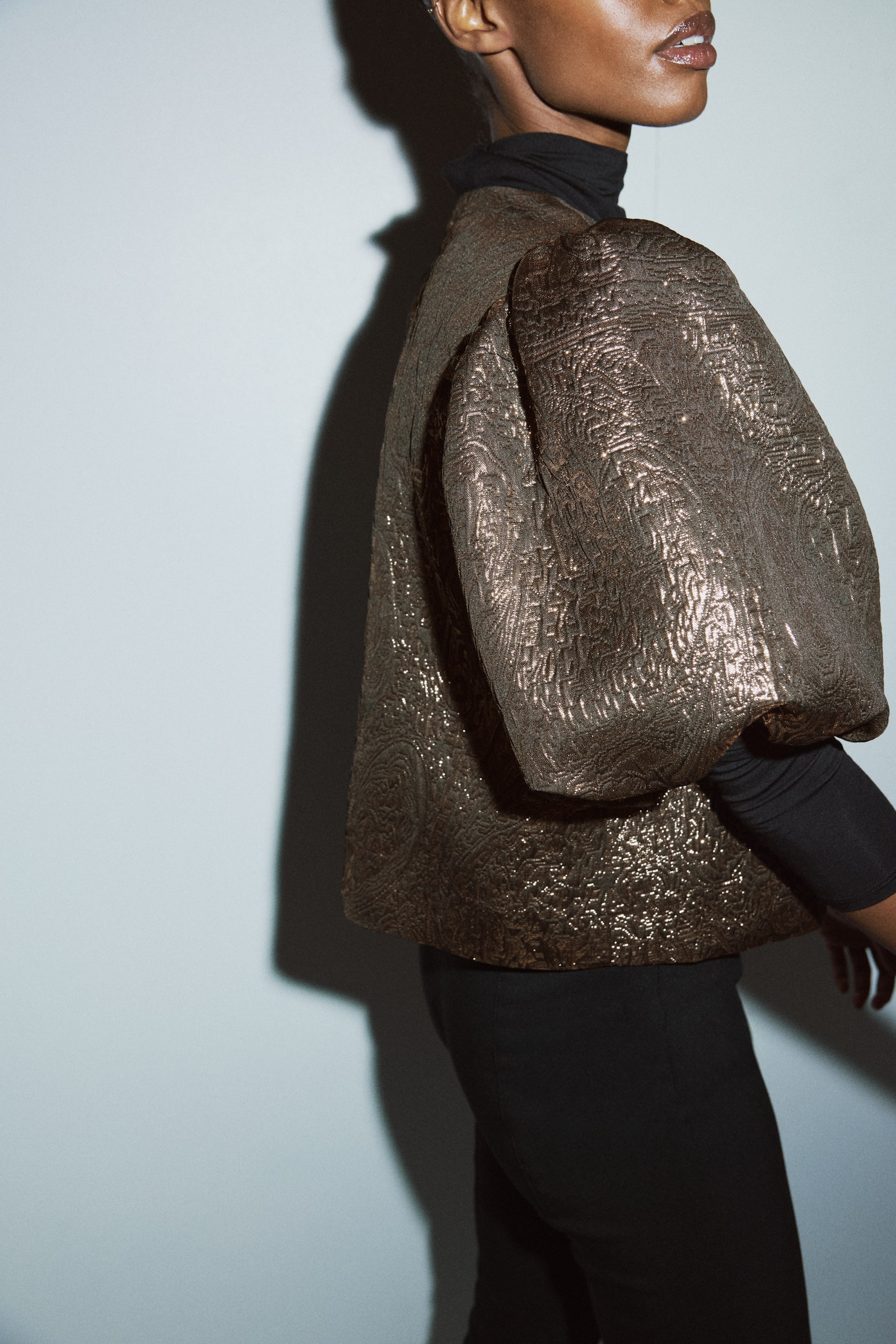 Zobacz większy obraz: Metaliczna kurtka w kolorze brązowo-złotym, widziana z profilu bocznego, posiada teksturowany, tłoczony wzór oraz obszerne rękawy 3/4 typu puff i jest noszona na czarnym golfie.