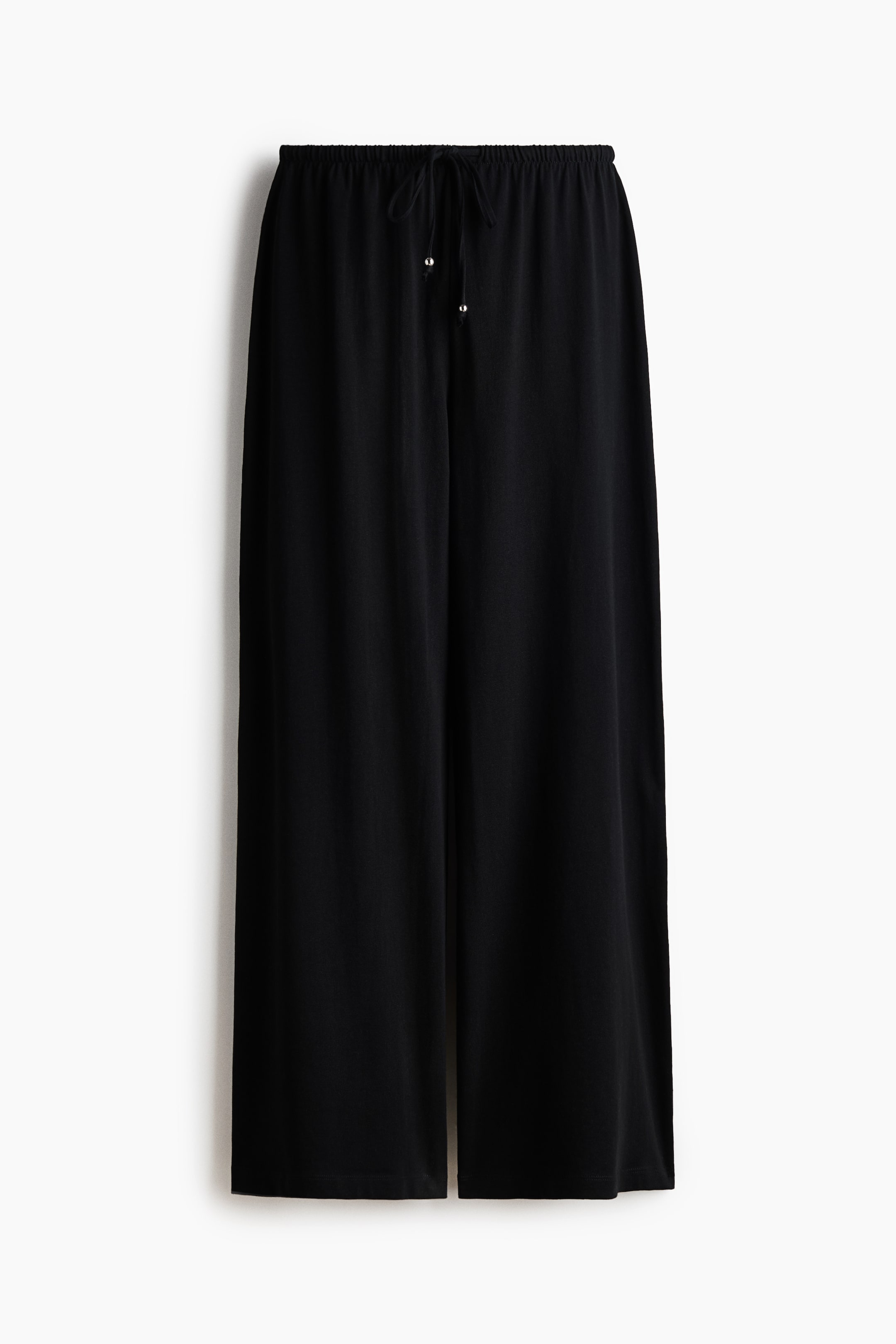 View larger image: Loose-fit trousers in black soft cotton jersey are showcased from the front. They feature an elasticated drawstring waist with a tied drawstring and silver-coloured metal tips, complemented by wide legs.