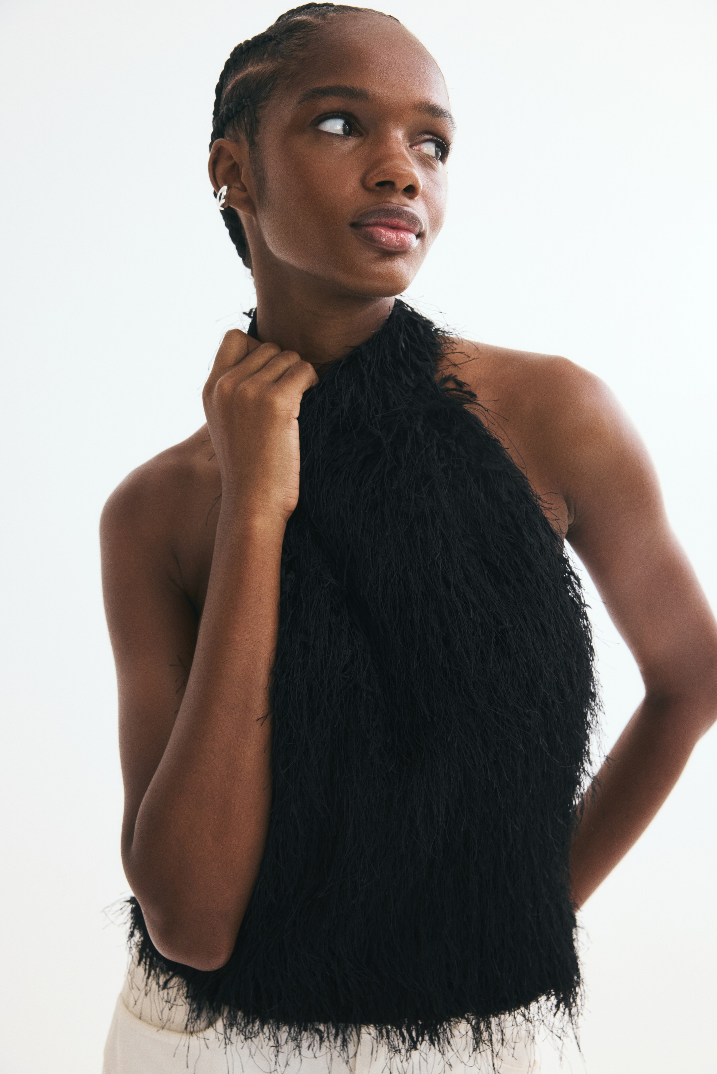 A black sleeveless top is shown from the front, featuring a high textured neckline and a slightly flared silhouette, fully covered in long, fine shaggy fringe resembling feathers.