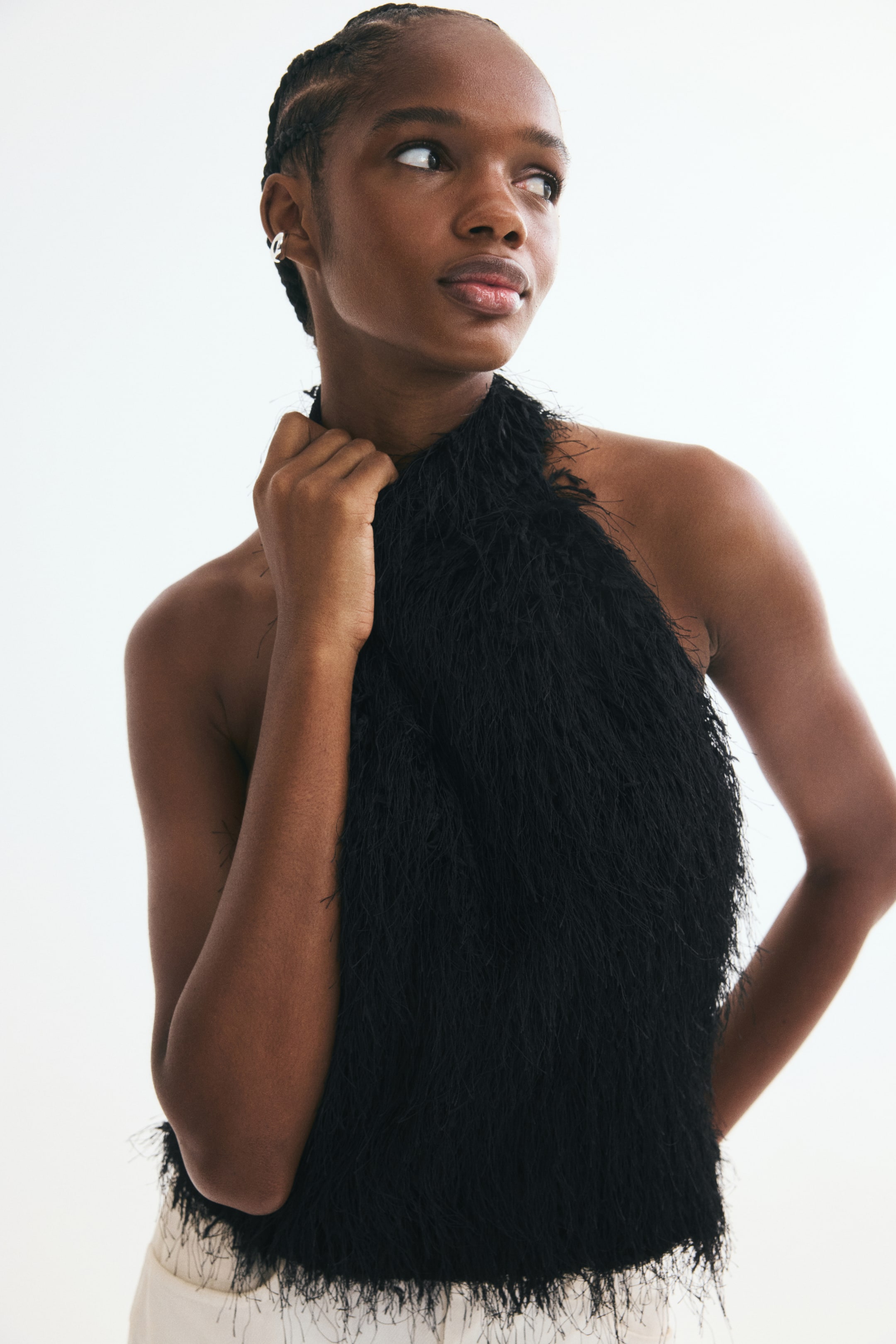 Größeres Bild ansehen: Ein schwarzes Neckholder-Top mit zotteliger, federartiger Textur wird von dem Model getragen, das mit einer Hand den hochgeschlossenen Ausschnitt berührt. Das ärmellose Top ist mit hellen Hosen kombiniert.