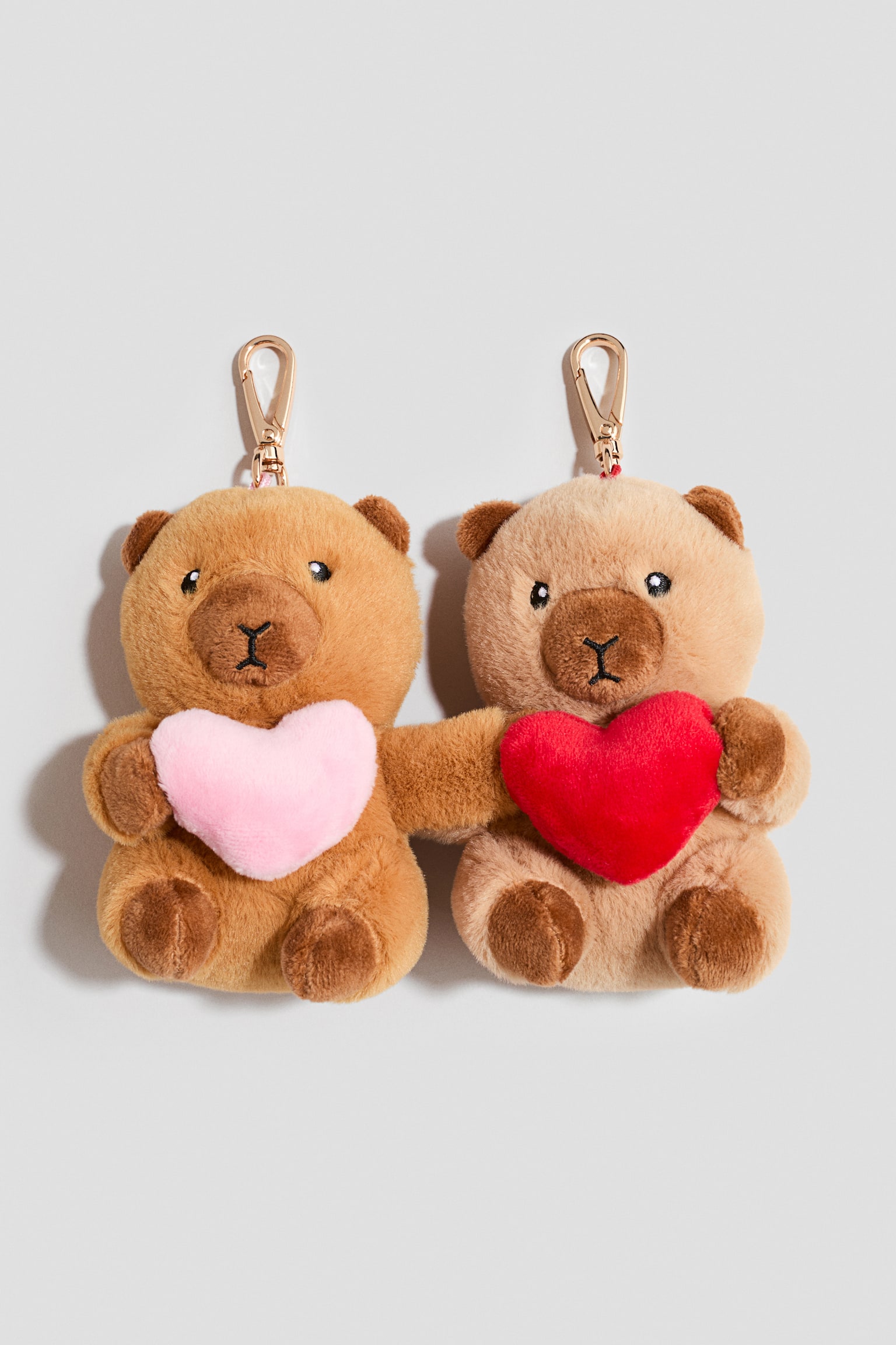 Zwei weiche Teddybär-Taschenanhänger sind nach vorne ausgerichtet, wobei ihre inneren Arme miteinander verbunden sind. Der goldbraune Bär links hält ein hellrosa Plüschherz, während der hellbeige Bär rechts ein rotes Plüschherz hält. Beide Anhänger verfügen oben über goldfarbene Metall-Karabinerverschlüsse.