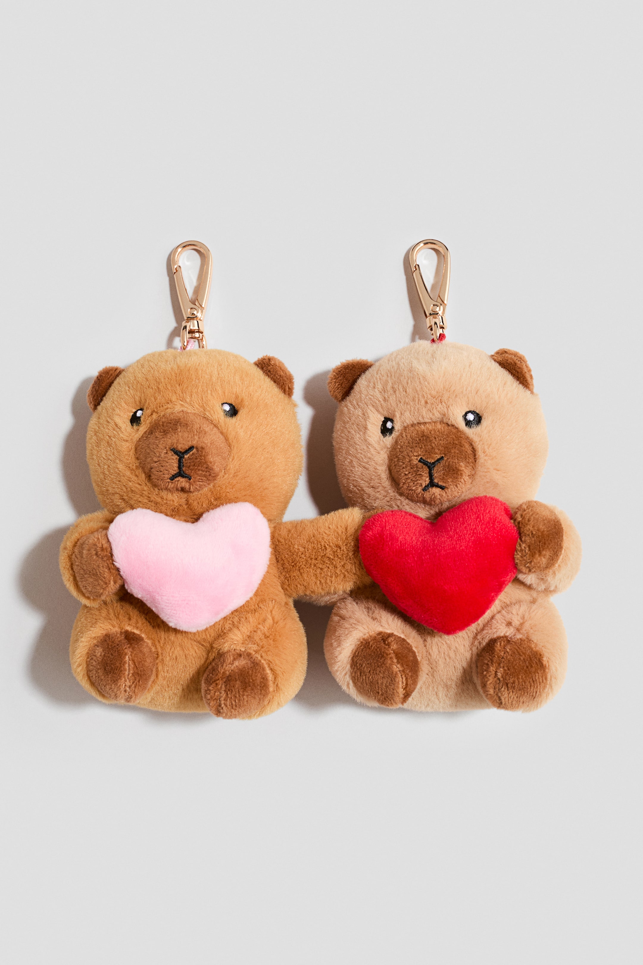 View larger image: Two plush teddy bear bag charms are facing forward, with their inner arms connecting them. The golden brown bear on the left is holding a light pink plush heart, while the light beige bear on the right is holding a red plush heart, with both charms featuring gold-toned metal lobster clasps at the top.