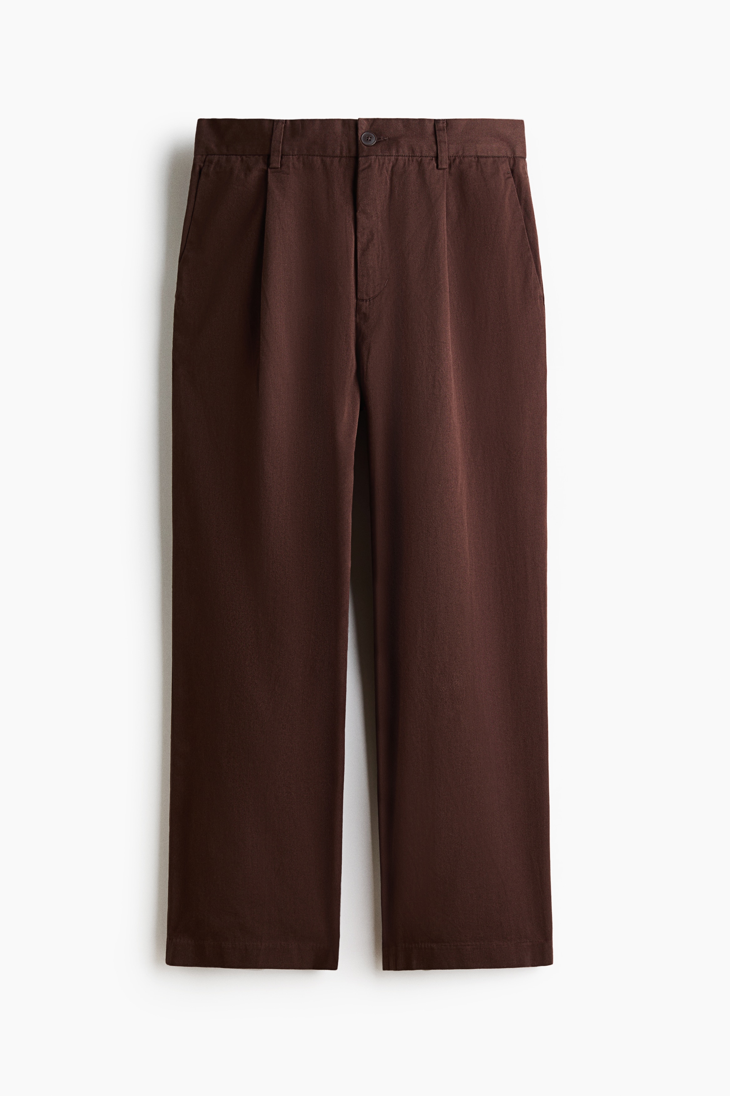 Brown cotton twill pants featuring a relaxed fit, straight legs, and subtle front pleats are being worn by the model. The pants are styled with an open, dark purple button-up shirt over a dark brown T-shirt and black sneakers.