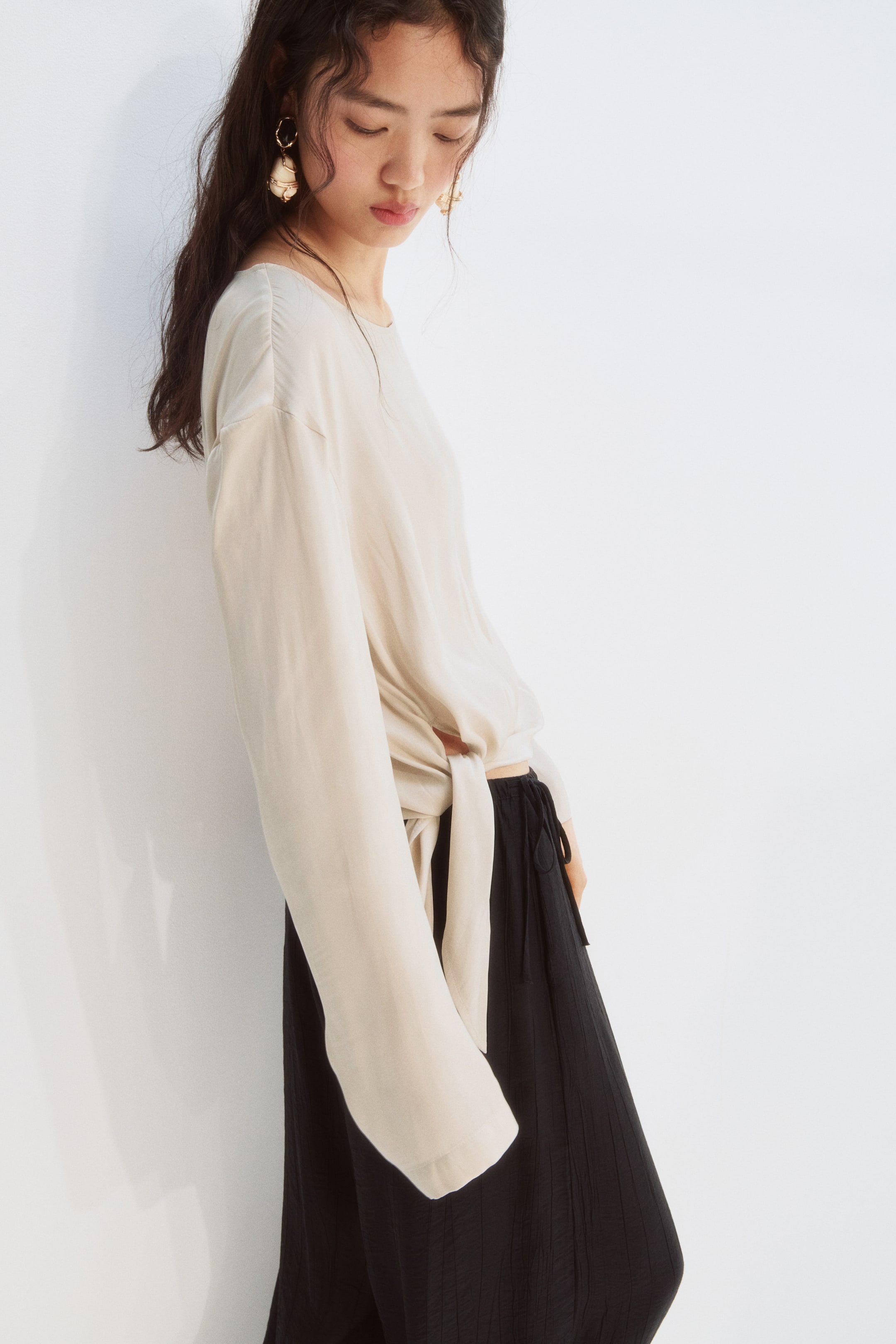 Agrandir l'image: Un haut beige clair à manches longues présente un col rond et une coupe ample, se nouant à la taille. Il est assorti à un pantalon noir à jambes larges avec une texture verticale subtile et une taille à cordon de serrage.