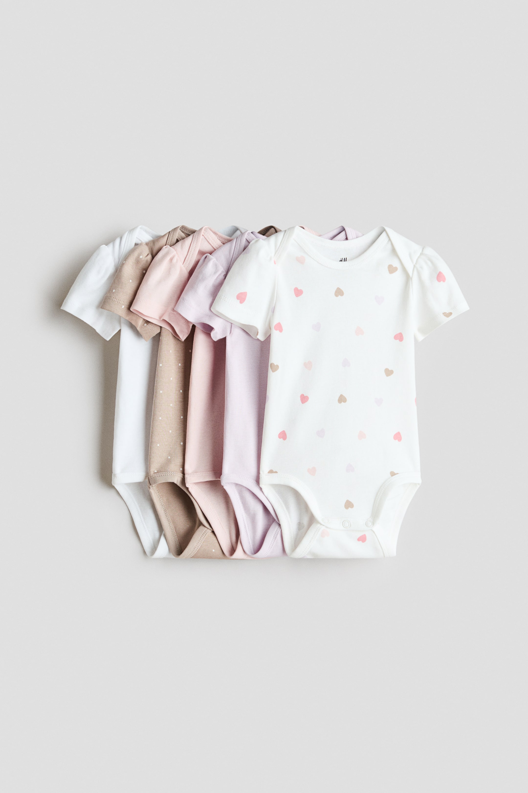 拡大画像を表示: 5枚の半袖ボディスーツが前面に部分的に重なり合って展示されています。肩には重ね縫いが施され、股下にはスナップボタンが付いています。前面のボディスーツは白地にピンクとブラウンのハート柄があり、他のものは様々なパステルカラーの無地と、白い水玉模様の薄茶色です。
