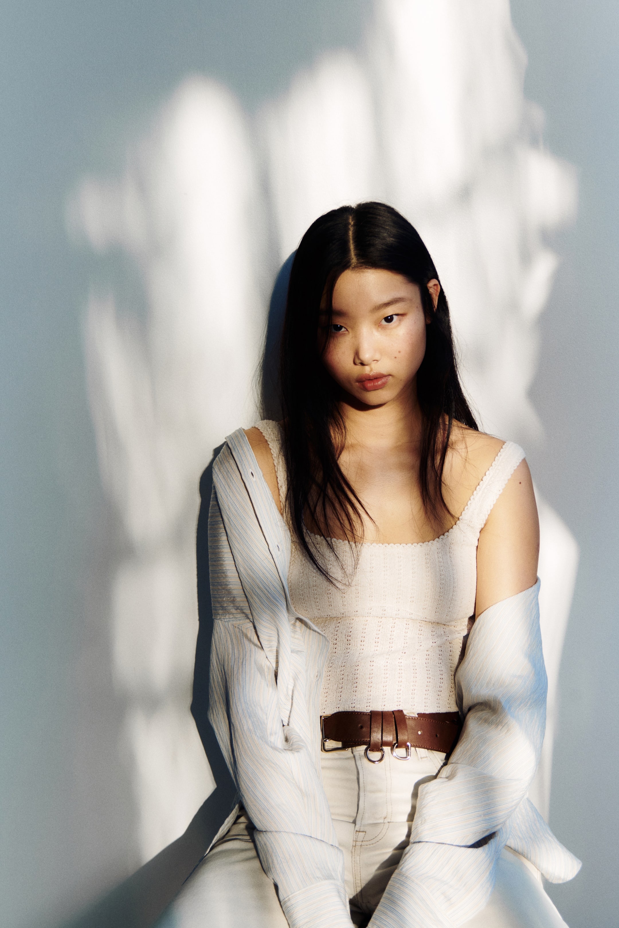 View larger image: "A cream open-knit camisole featuring a square neckline and scalloped trim is being worn. A light blue striped button-down shirt is casually draped open over the shoulders. A dark brown leather belt with a double metal buckle elegantly cinches the waist over cream-coloured trousers."