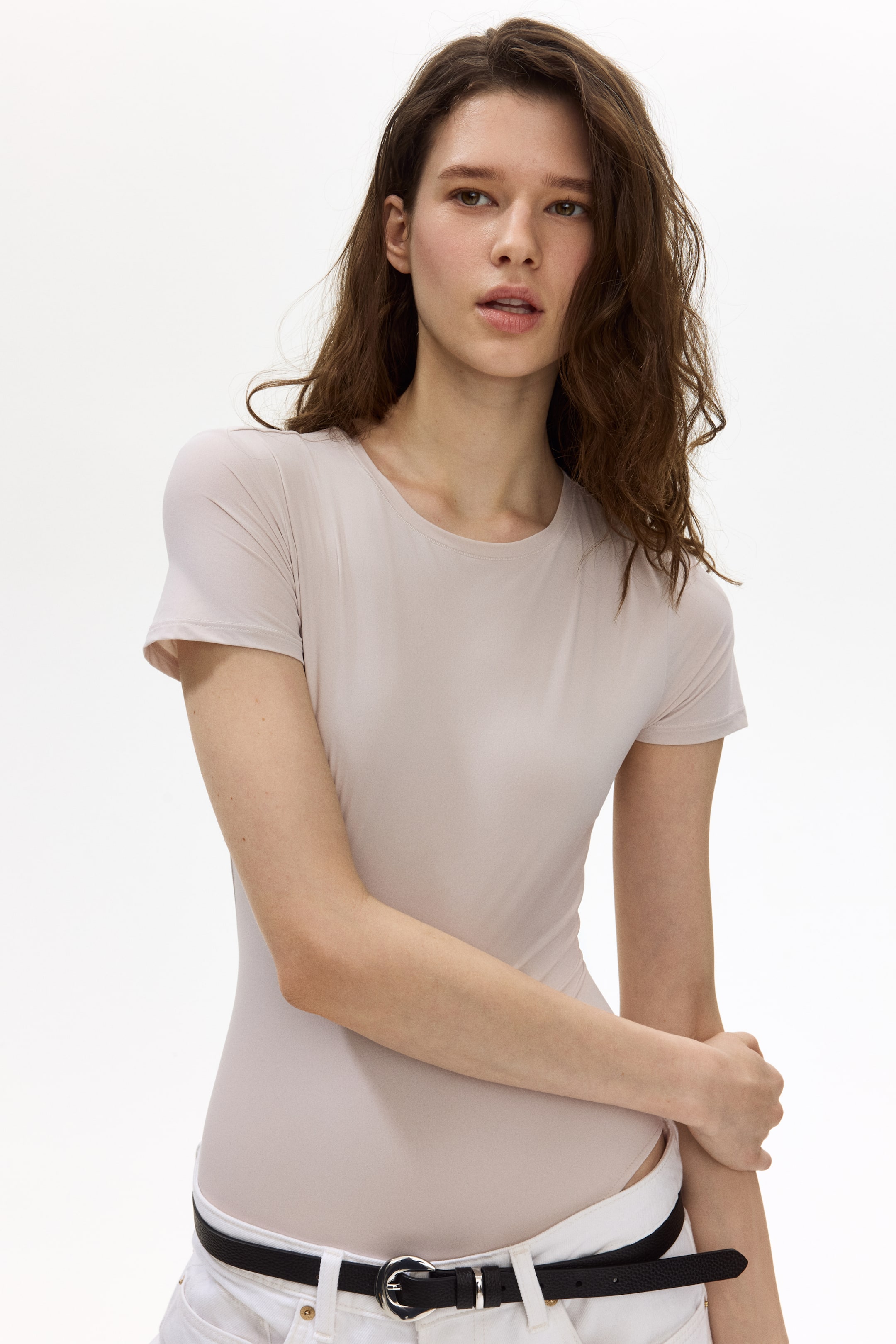 Grotere afbeelding bekijken: Een licht beige, aansluitende bodysuit met een ronde hals en korte mouwen, waarbij de gladde stof lijkt ingestopt in witte broek en vastgezet met een zwarte riem.