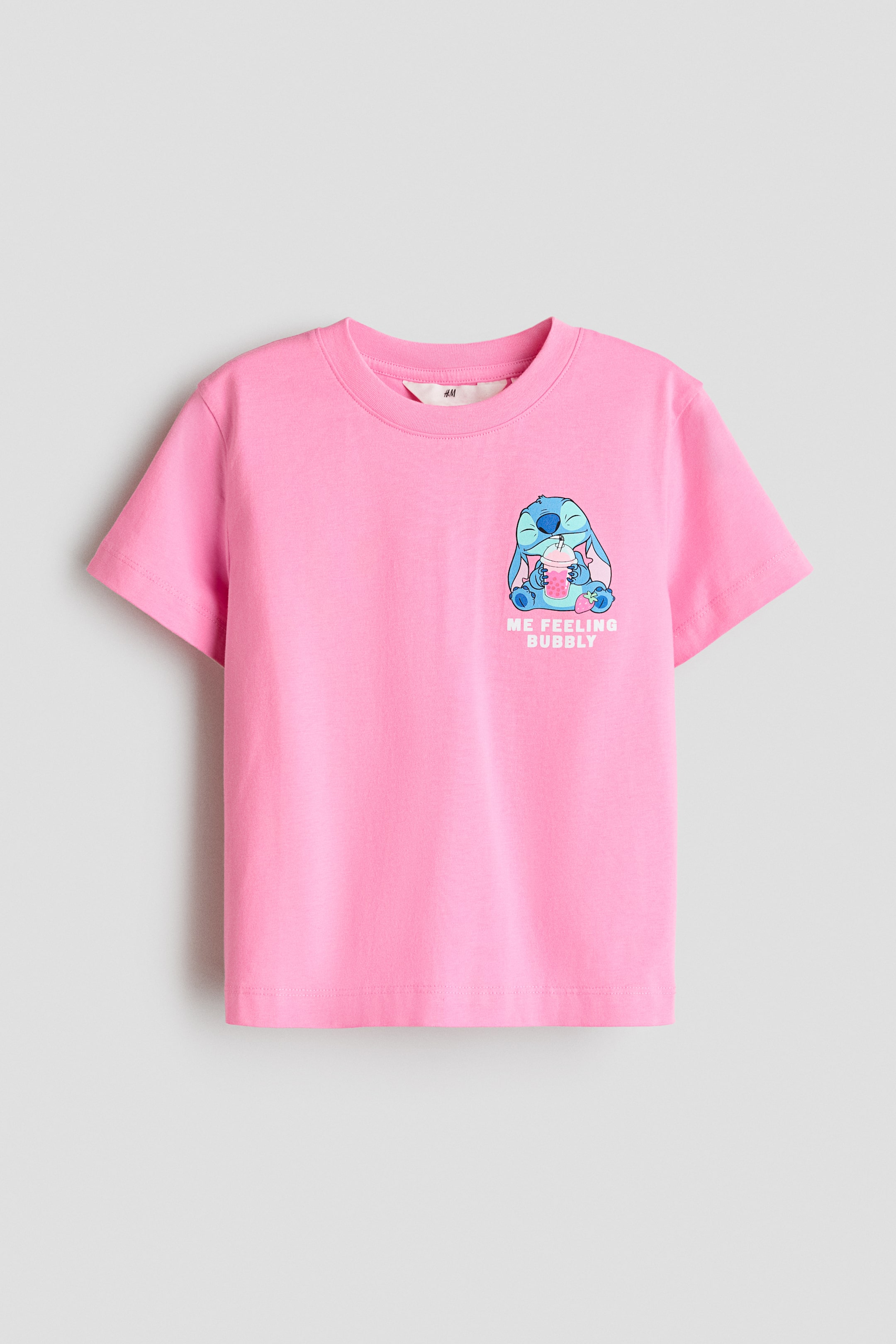 Visualizza immagine più grande: Una t-shirt rosa mostra il suo fronte, caratterizzata da uno scollo rotondo e maniche corte. Un motivo stampato colorato sul davanti raffigura un personaggio alieno blu che tiene in mano una bevanda rosa con una cannuccia, circondato da piccoli frutti, e il testo bianco 'ME FEELING BUBBLY'.