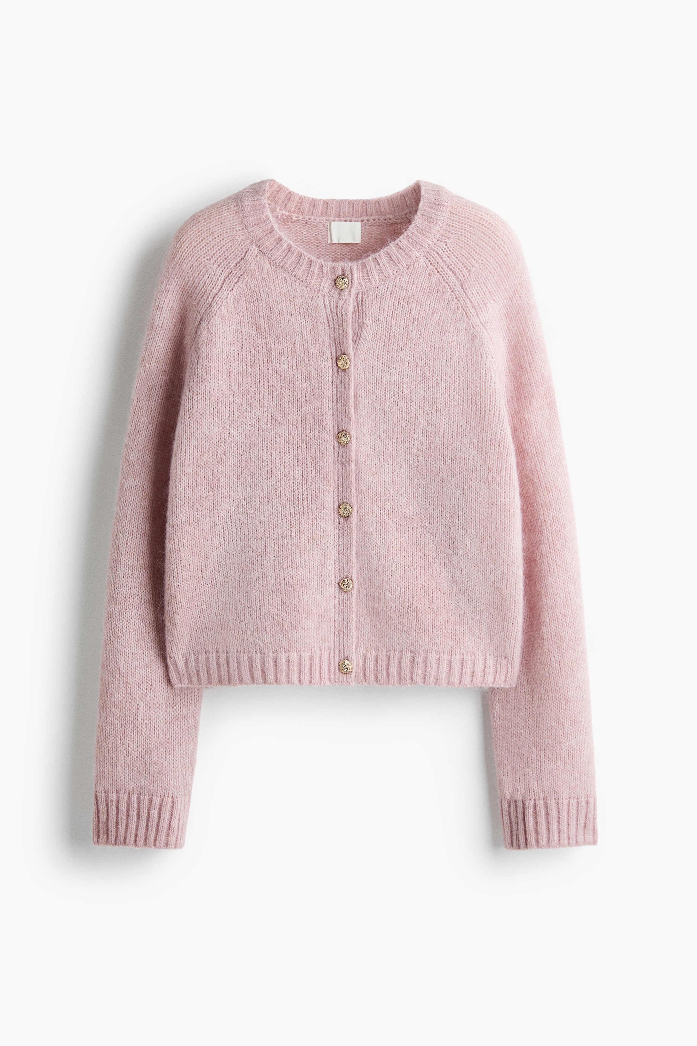 The model is wearing a light pink, soft-knit cardigan with a round ribbed neckline and textured gold buttons. The oversized cardigan features long raglan sleeves and a ribbed hem, and is styled partially unbuttoned over dark-wash jeans.