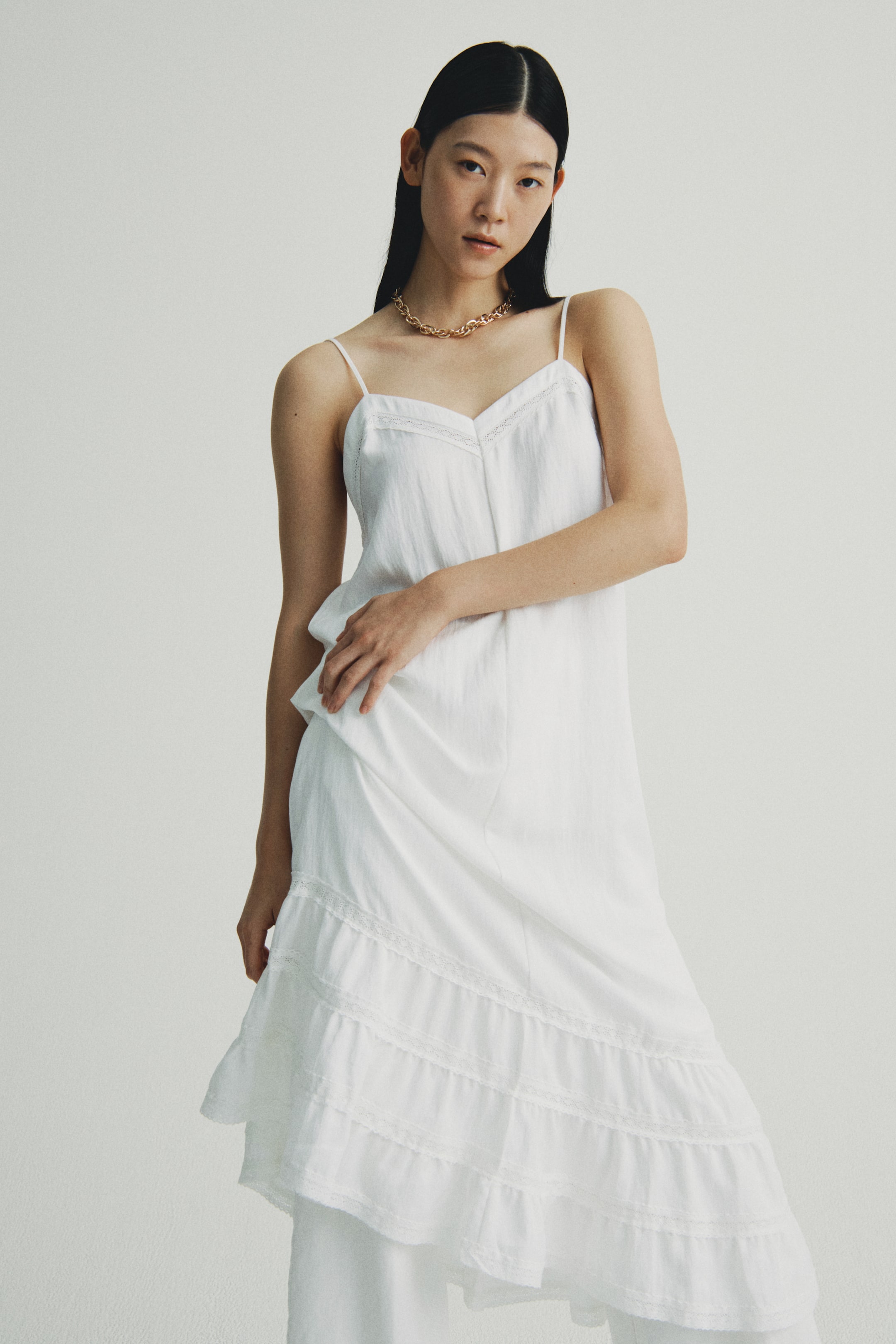 Xem ảnh lớn hơn: Một chiếc áo không tay có kết cấu màu trắng và chân váy xếp tầng đang được trình bày từ góc nhìn phía trước hơi nghiêng về bên phải. Chiếc áo có dây spaghetti và cổ chữ V với viền tinh tế, trong khi chiếc váy có nhiều tầng xếp nếp với chi tiết ren và được mặc bên trên quần trắng.
