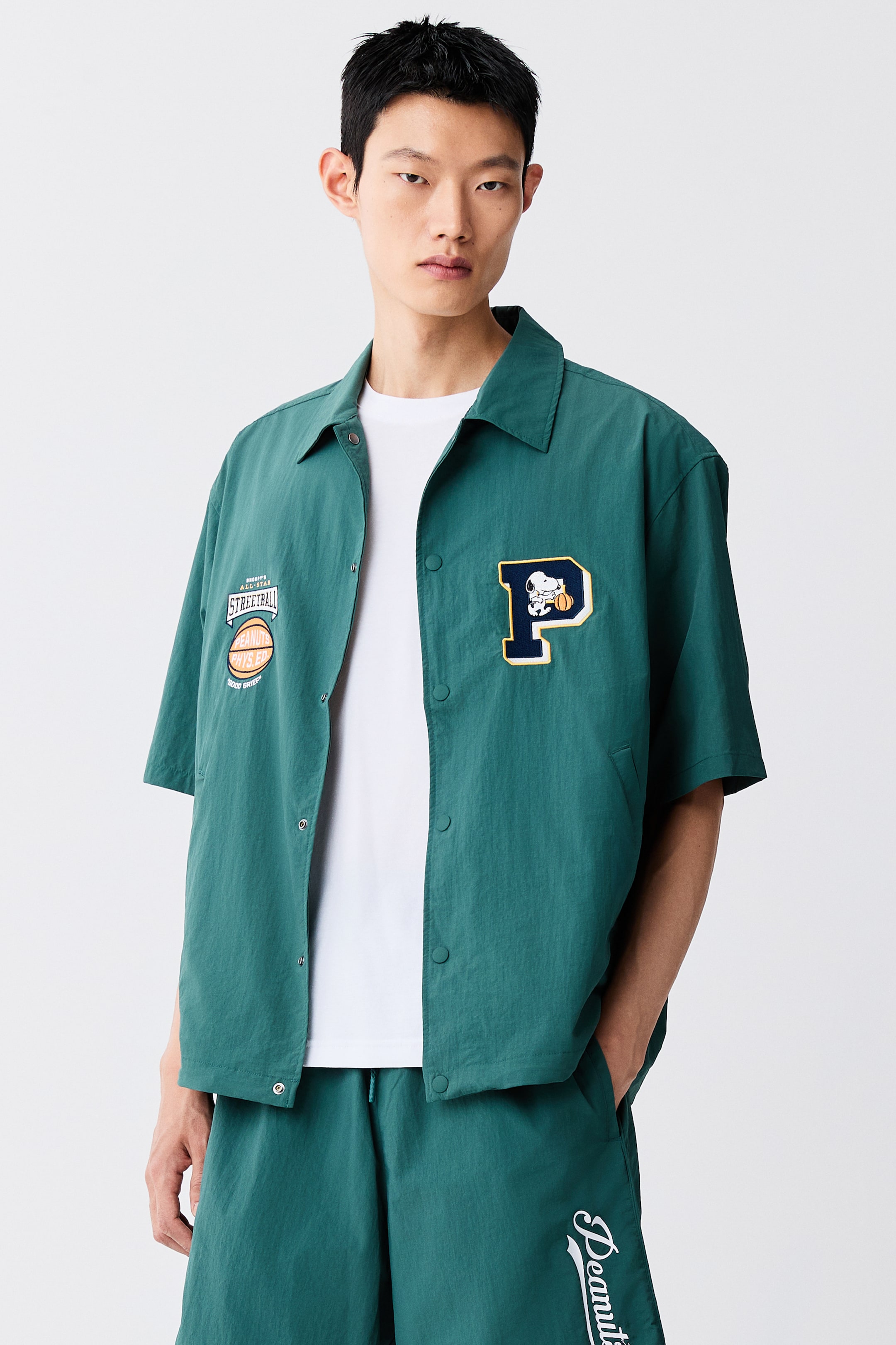 View larger image: A short-sleeved green coach jacket is worn open by the model over a white t-shirt and matching green shorts. The jacket features a classic collar, snap-button closures, and embroidered patches on the chest, including a large letter "P" motif.