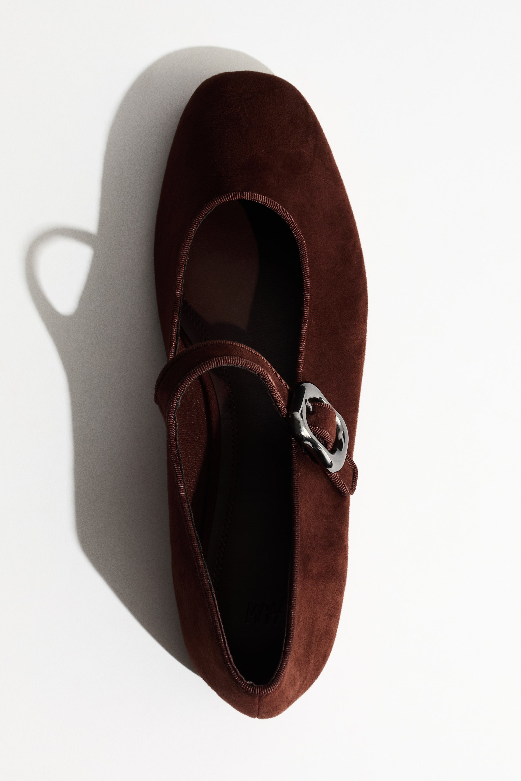 Ver imagen más grande: Un zapato plano tipo Mary Jane de color marrón oscuro se presenta desde un ángulo cenital, mostrando una textura similar al ante. Una correa cruza el empeine, asegurada por una hebilla oscura de forma irregular.