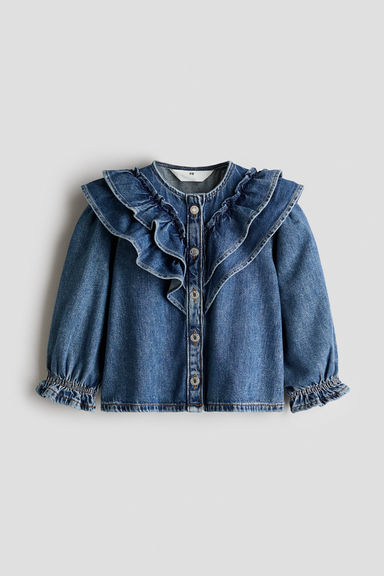 A long-sleeved denim blouse in a medium wash is shown from the front, featuring a large ruffled collar that drapes over the shoulders. The blouse has a front button-down fastening with metal buttons and long sleeves with elasticated, ruffled cuffs.