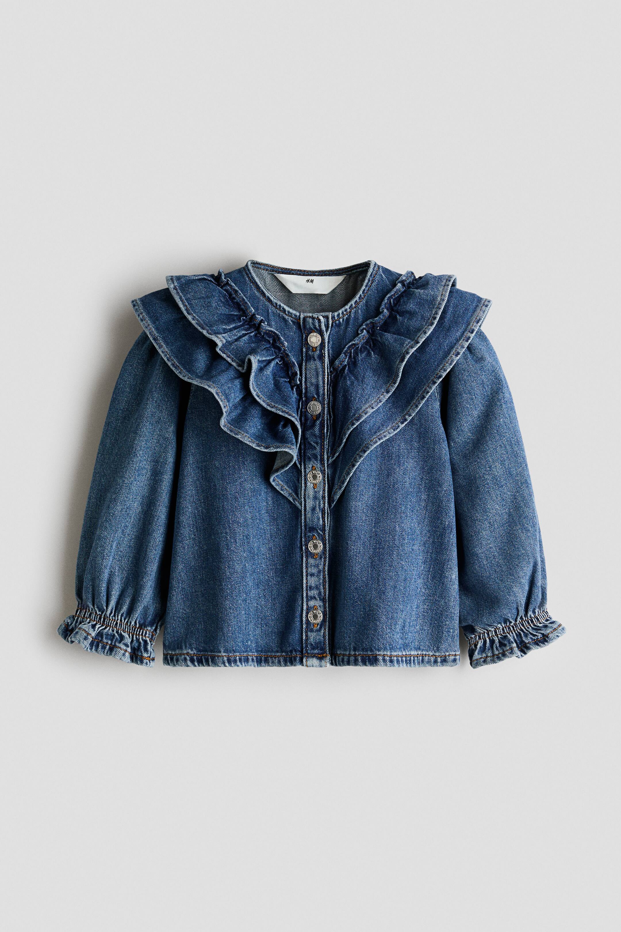 Büyük resmi görüntüle: Orta yıkamalı, uzun kollu denim bluz, önden görünümde olup omuzların üzerine dökülen büyük fırfırlı bir yakayı sergiliyor. Bluz, metal düğmeli ön düğme kapaması ve elastik, fırfırlı manşetlere sahip uzun kollara sahiptir.
