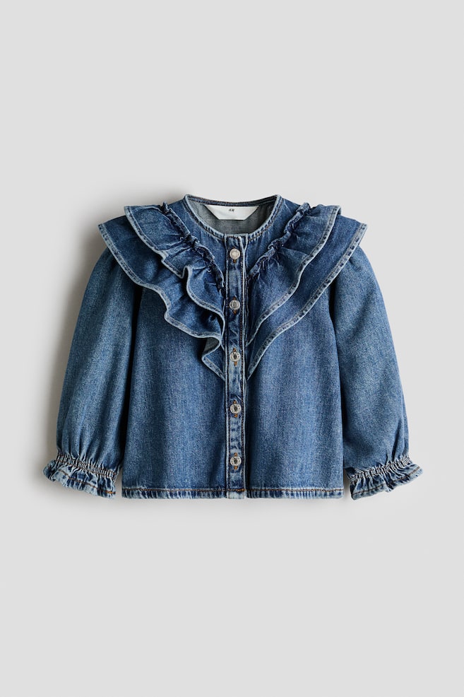 A long-sleeved denim blouse in a medium wash is shown from the front, featuring a large ruffled collar that drapes over the shoulders. The blouse has a front button-down fastening with metal buttons and long sleeves with elasticated, ruffled cuffs. - 1
