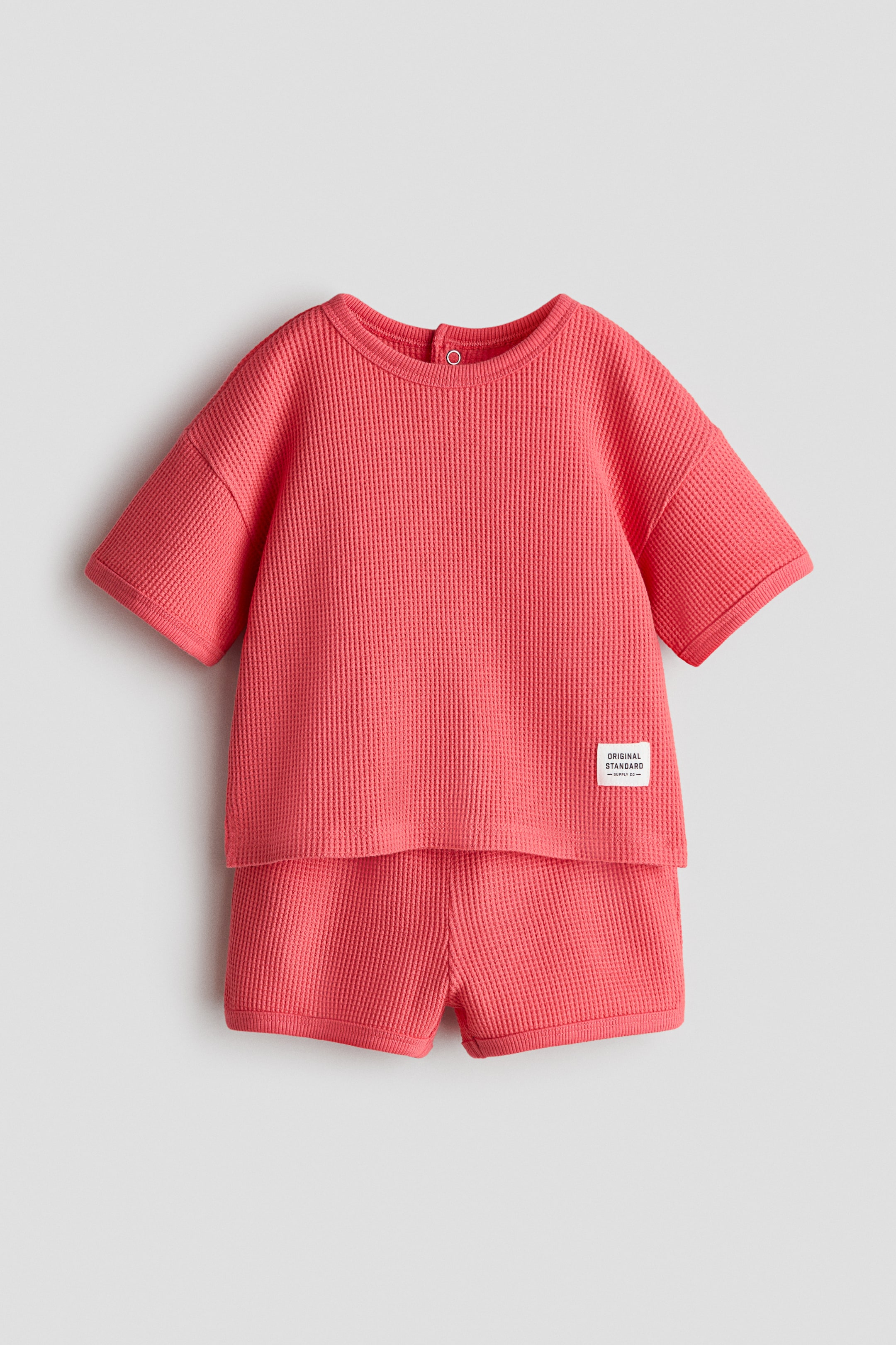 View larger image: Introducing from the front, a coral waffled jersey t-shirt and shorts set features a t-shirt with a round, ribbed neckline, a press-stud visible at the back neckline, dropped shoulders, rib-trimmed cuffs, and a white woven label on the lower right front. The matching shorts showcase a smooth waistline and rib-trimmed leg openings.