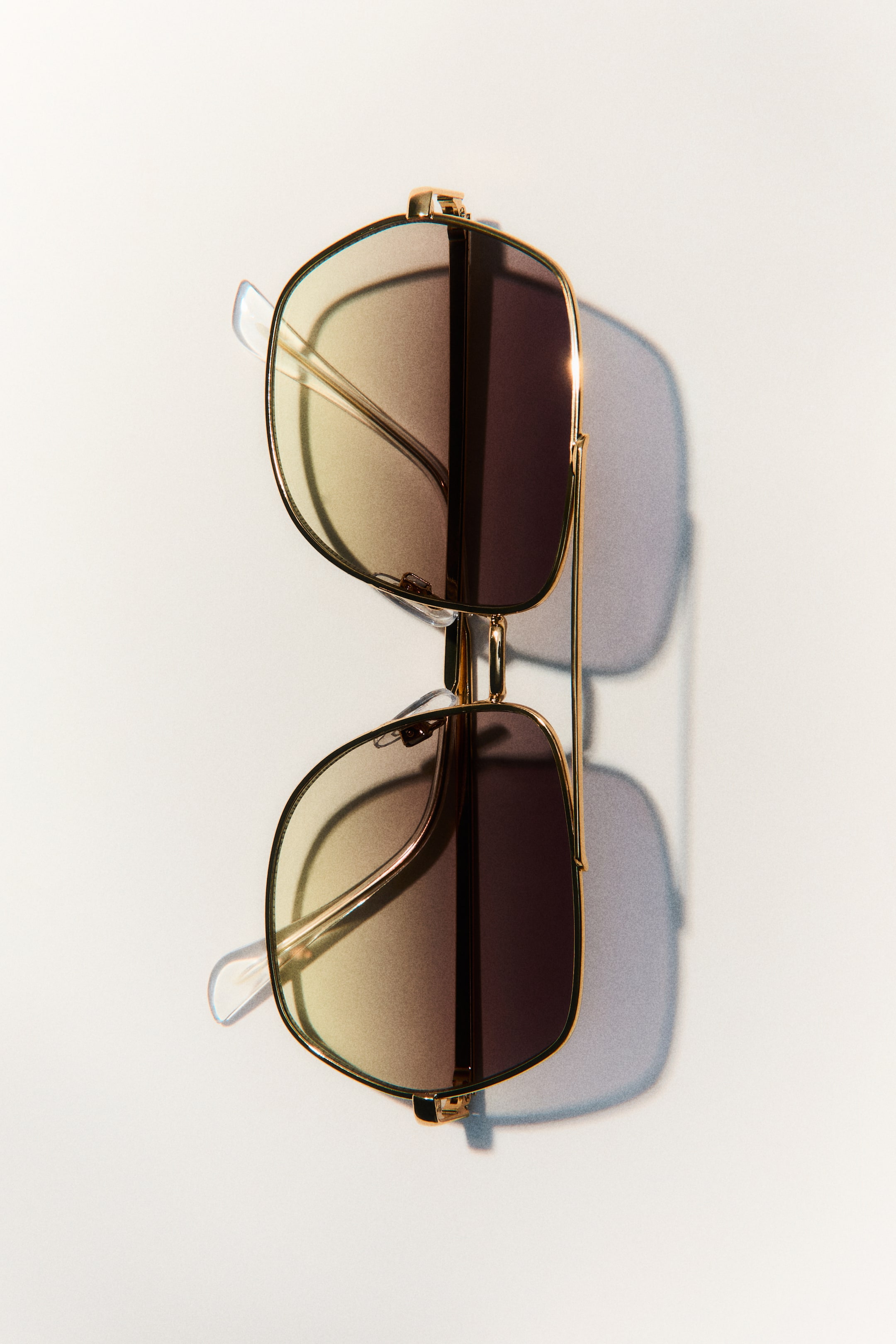 View larger image: Sunglasses are laid flat, positioned vertically, showcasing a gold-toned double bridge frame with gradient lenses that shift from light yellow-brown to dark brown. Clear nose pads and temple tips are visible.