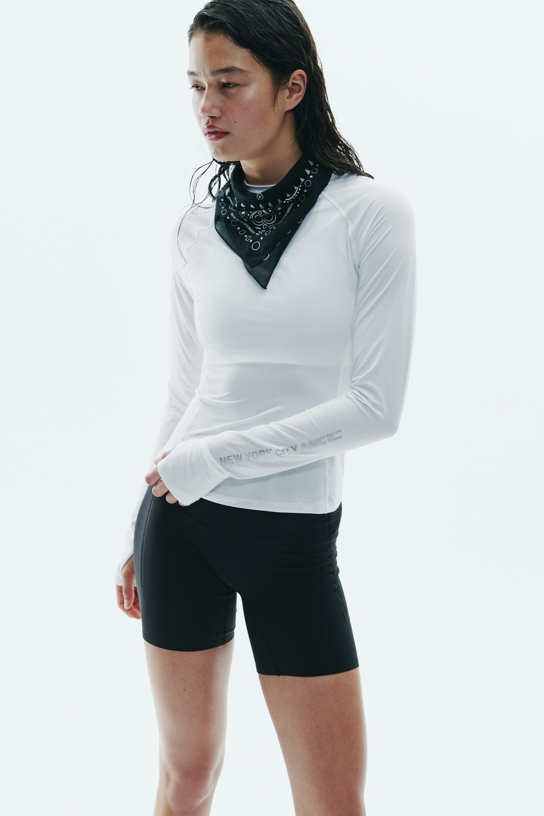 Ampliar la imagen: La modelo lleva puesta una playera atlética blanca de manga larga con mangas raglán y texto plateado impreso en el antebrazo. La prenda se combina con un bandana negro estampado atado al cuello y shorts atléticos negros ajustados.
