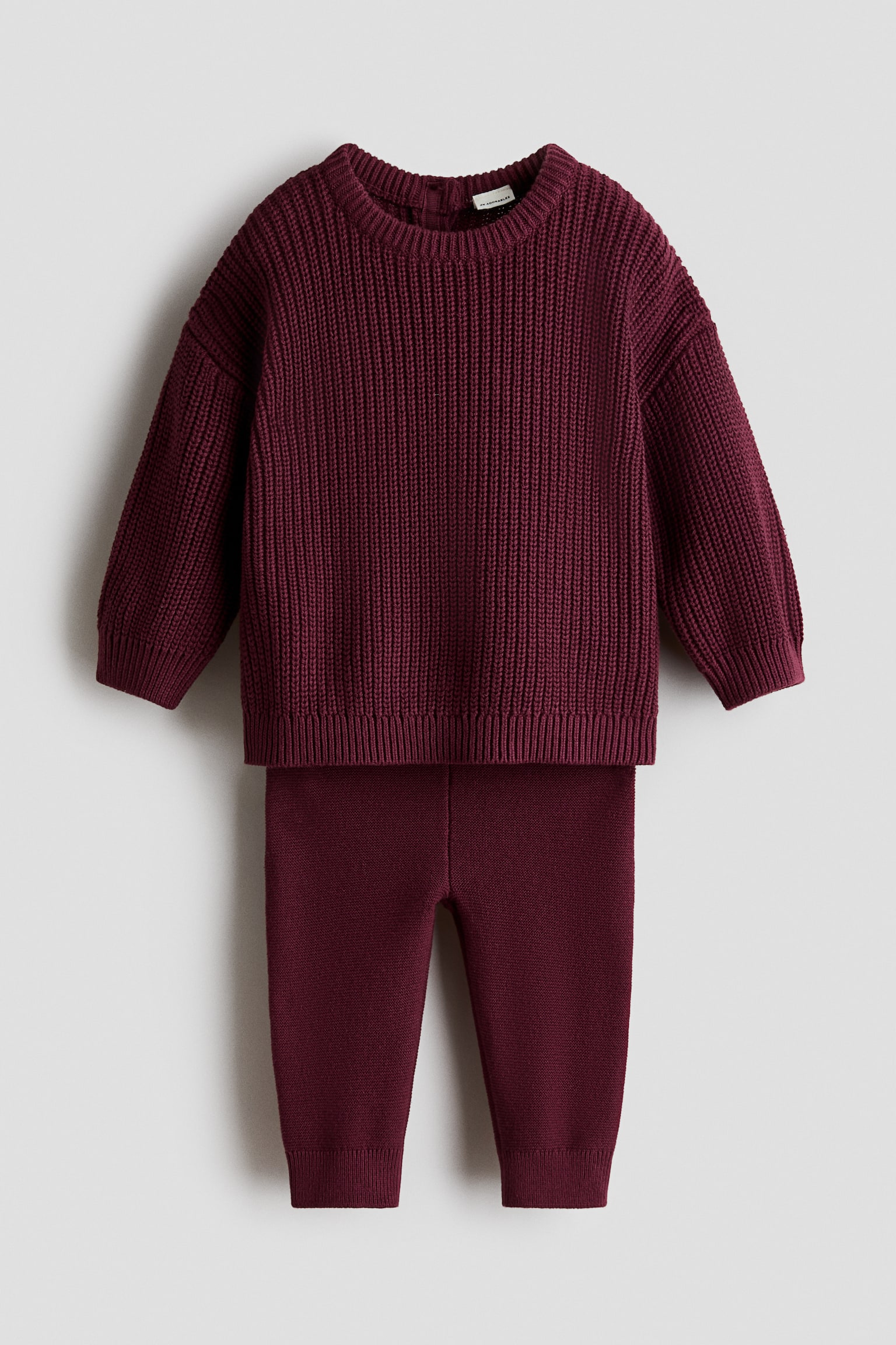 A burgundy two-piece knit set is displayed from a front-facing angle, featuring a patent-knit sweater with a round neckline, dropped shoulders, and ribbed trim. The matching fine-knit leggings feature ribbed ankle cuffs. - 1