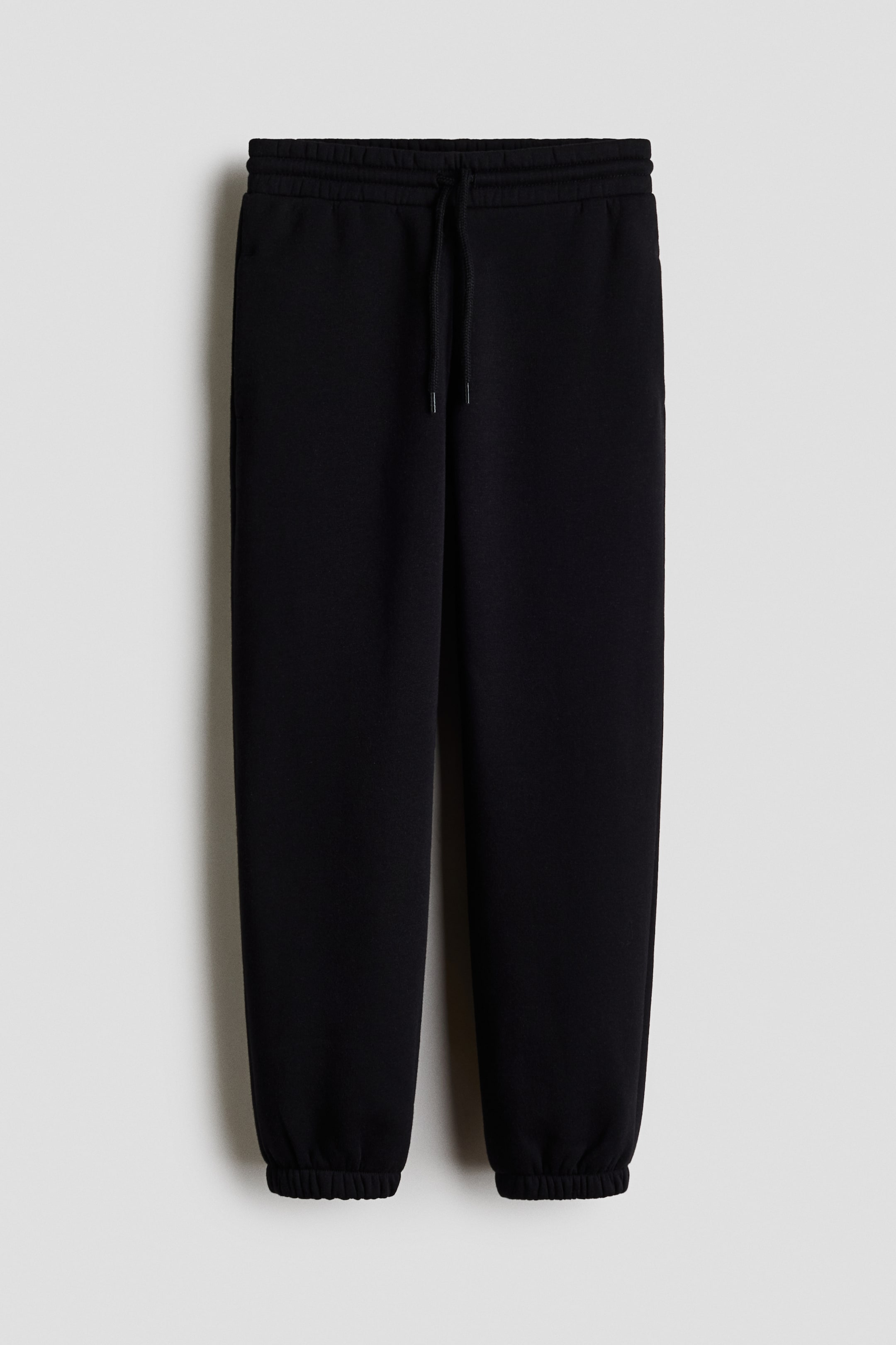 View larger image: "Black joggers in sweatshirt fabric are being shown from the front, featuring an elasticated drawstring waist, subtle side pockets, and covered elastication at the hems."