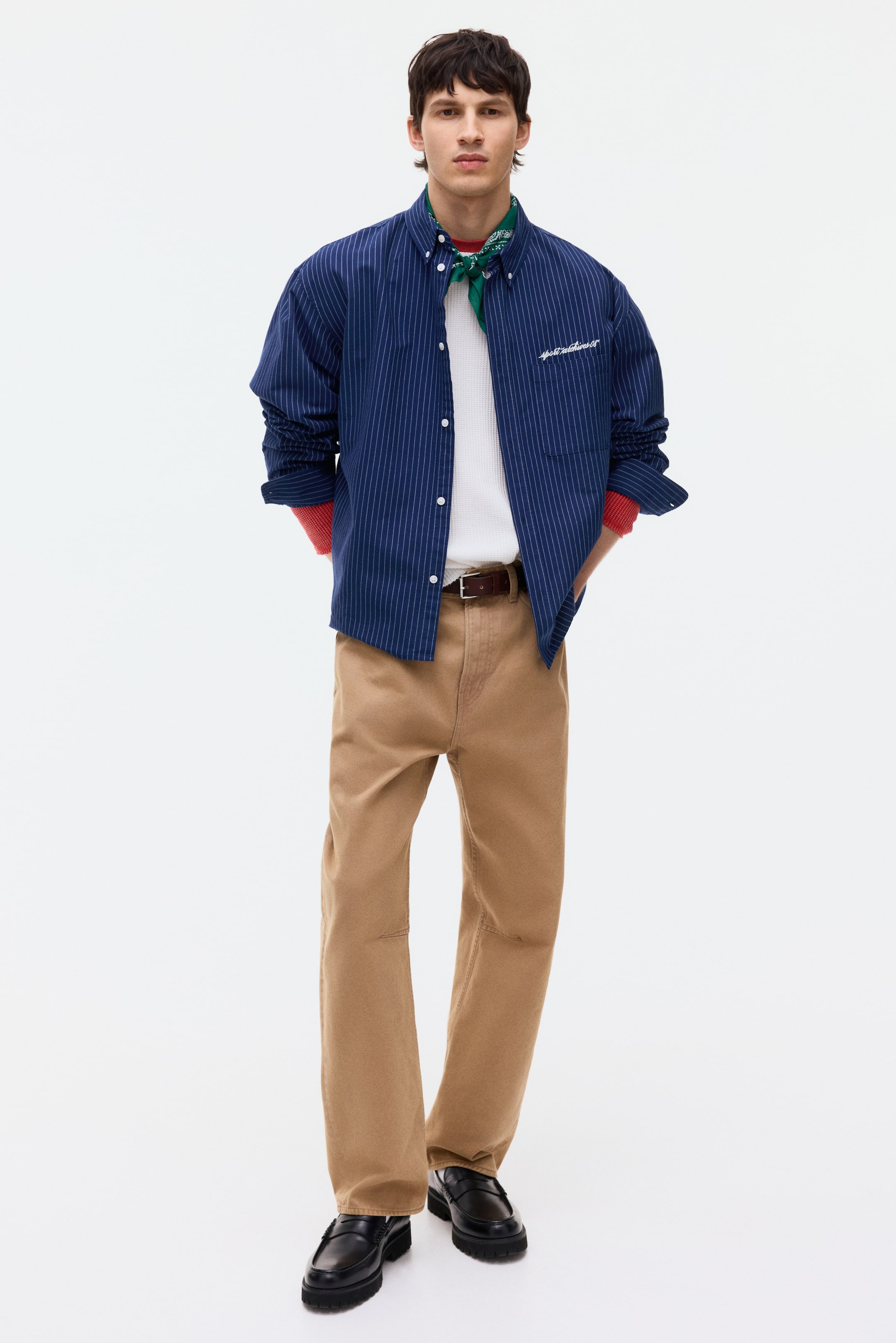 הצגת תמונה גדולה יותר: חולצה כחולה עם פסים דקים וצווארון כפתורים, טקסט רקום וכיס פתוח בחזה נלבשת על ידי הדוגמן בגזרה רפויה, פתוחה מעל חולצה לבנה. השרוולים הארוכים של החולצה מגולגלים, וחושפים חולצה אדומה מתחת, ובנדנה ירוקה נראית בצווארון.