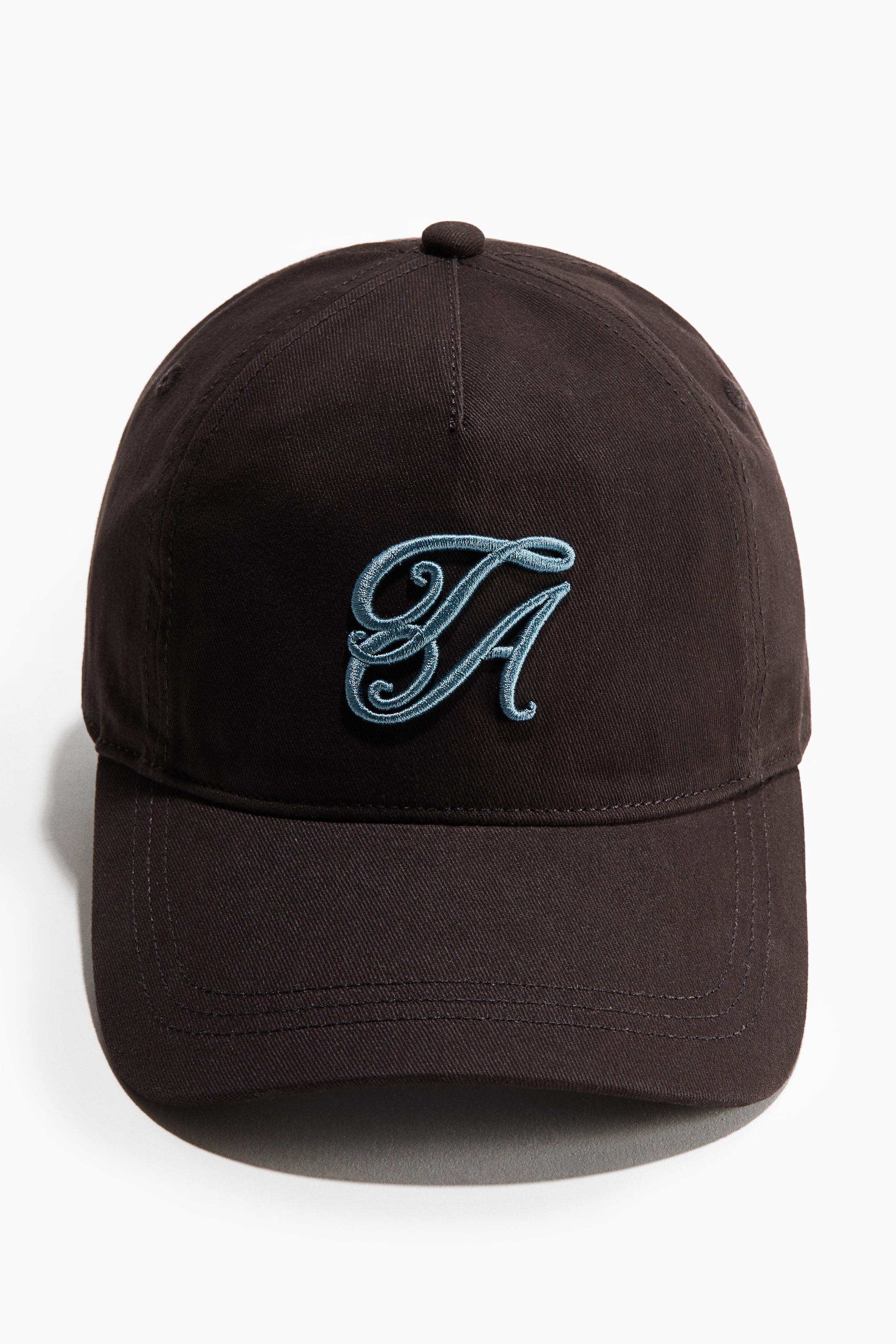 Ver imagen más grande: Se muestra una gorra de sarga de algodón marrón oscuro desde una vista frontal, con un monograma estilizado bordado en azul claro en su parte delantera. Presenta una visera curva y un botón en la parte superior de su corona.