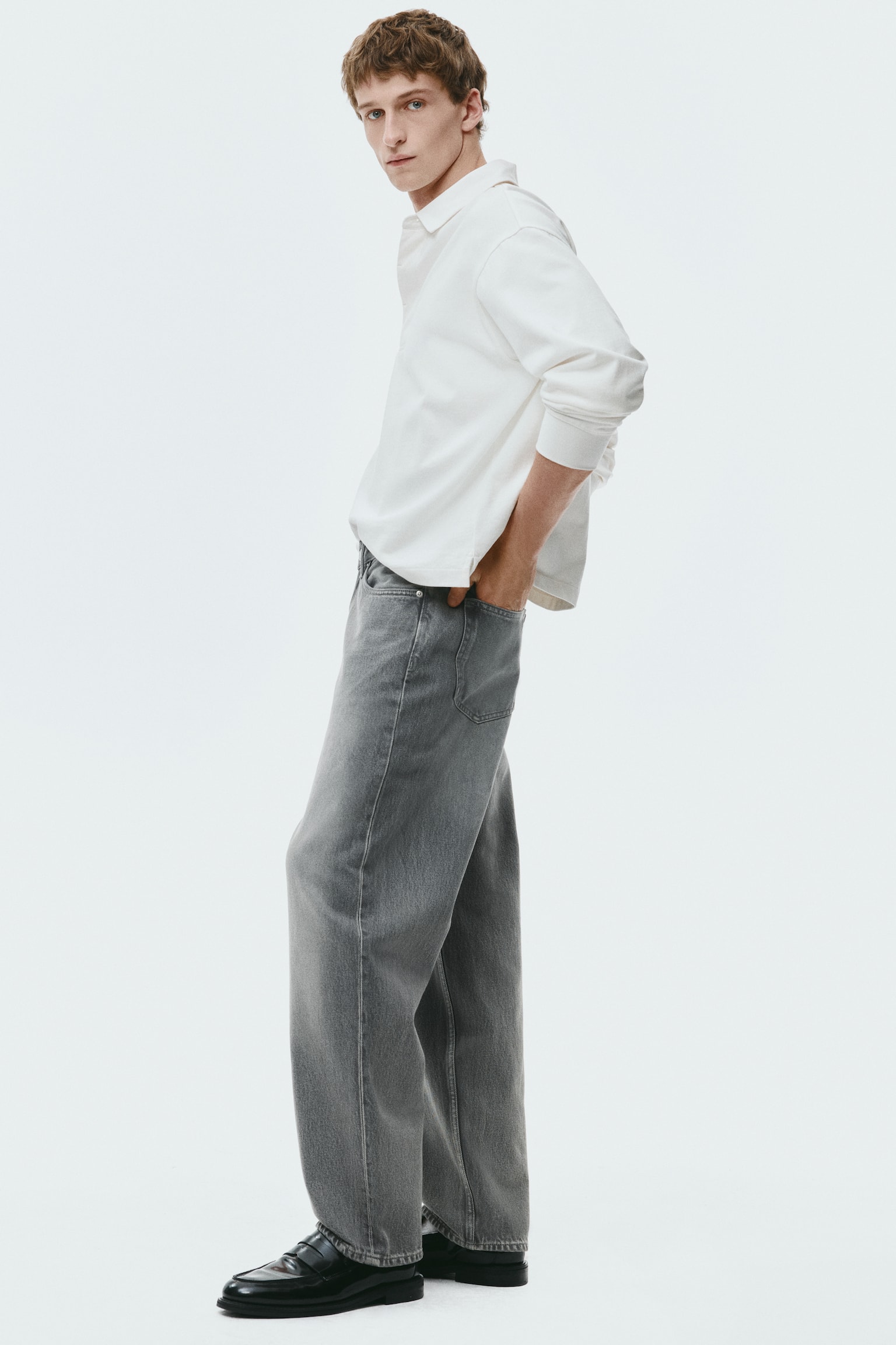 A white long-sleeved polo shirt with elasticated cuffs is being worn with loose-fitting, washed grey wide-leg jeans and black chunky-soled loafers, all presented at a slight right-facing angle. - 1