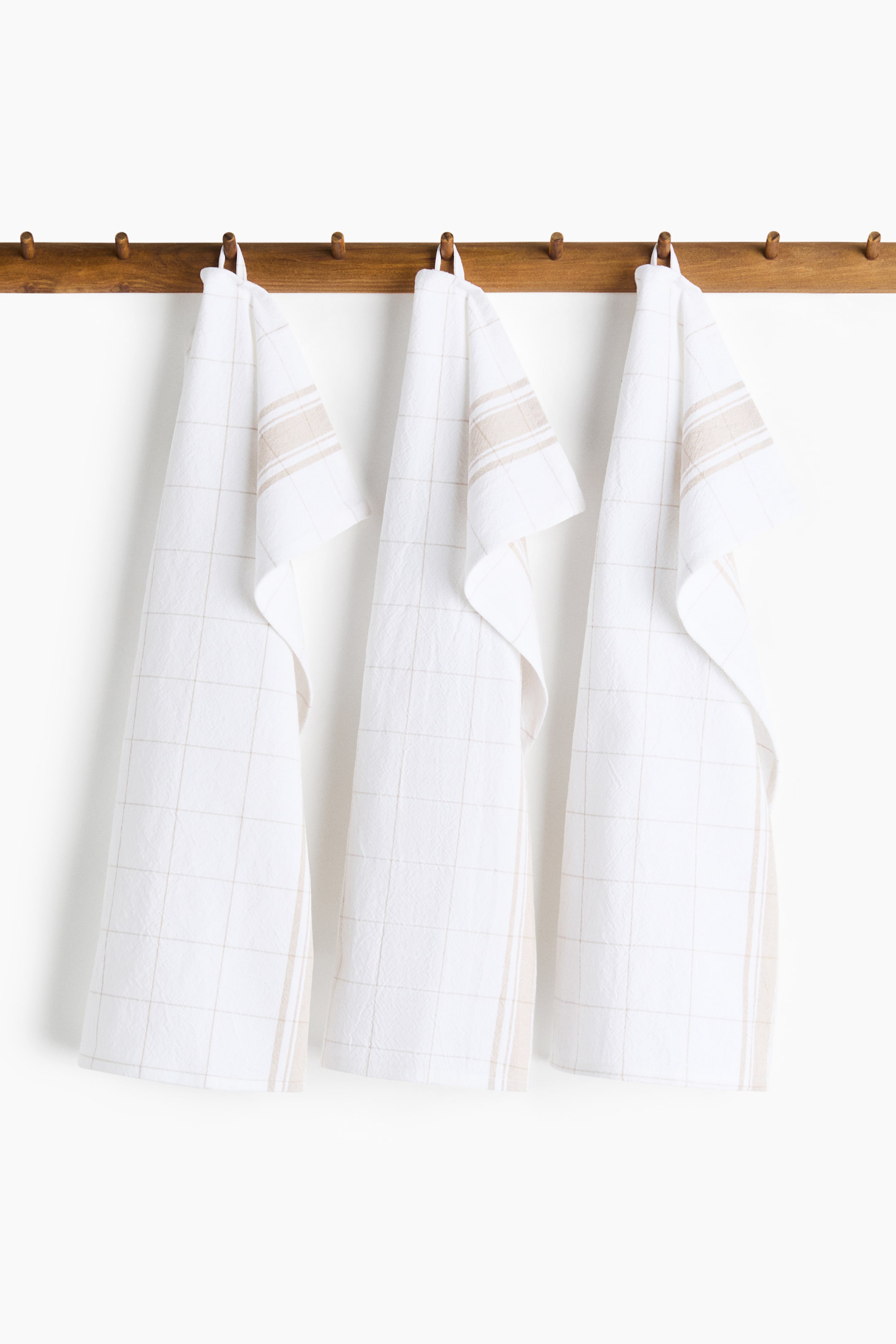 View larger image: Three cotton tea towels hang from a wooden rack, facing forward. Each white towel features a subtle grid pattern and beige stripes, and is designed with an integrated hanger on one short side.