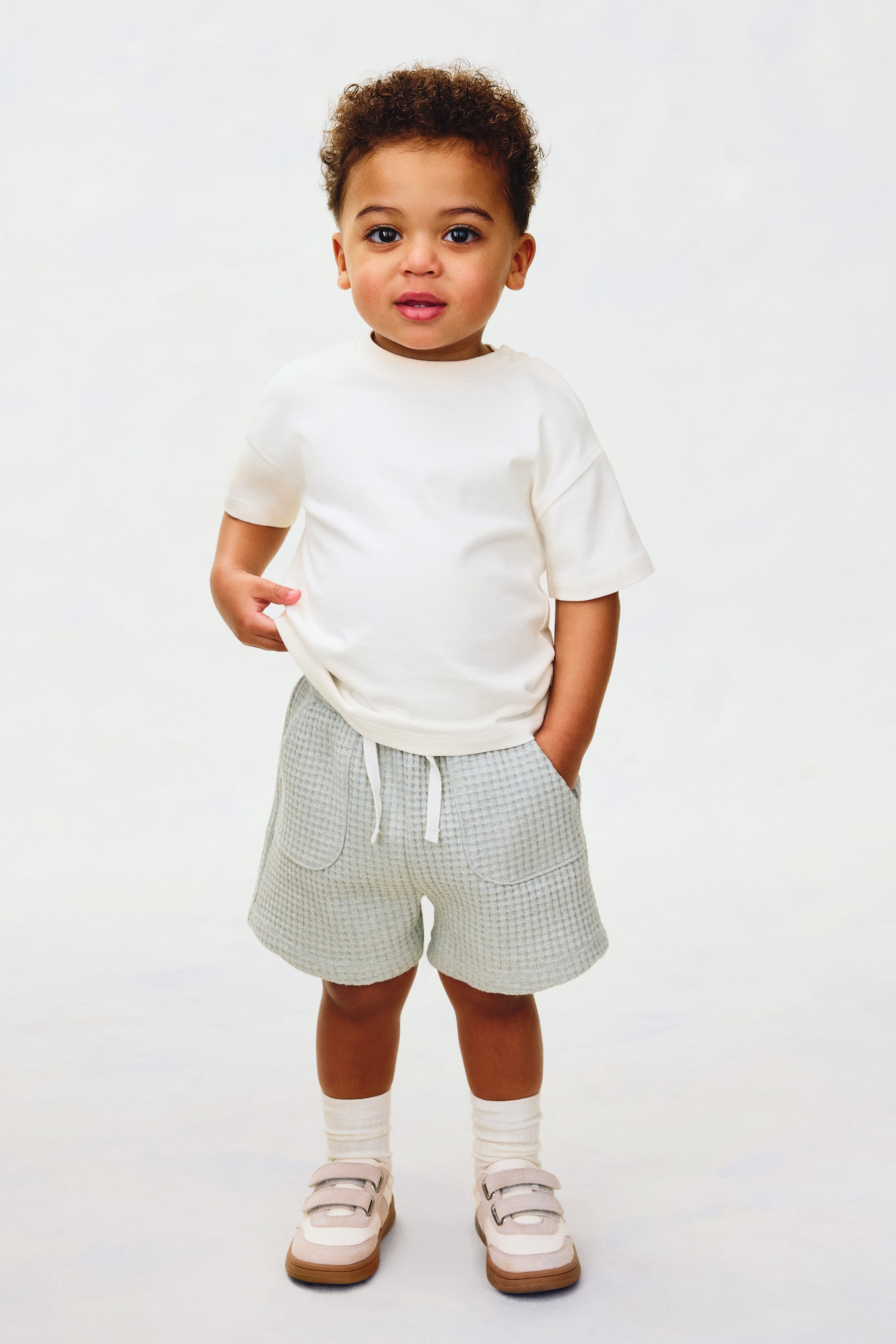 Grösseres Bild ansehen: Ein cremefarbenes T-Shirt mit kurzen Ärmeln präsentiert einen Rundhalsausschnitt und eine entspannte Passform. Hellgrüne Waffelstrick-Shorts verfügen über einen elastischen Kordelzugbund mit weißen Bändern und zwei aufgesetzten Vordertaschen.