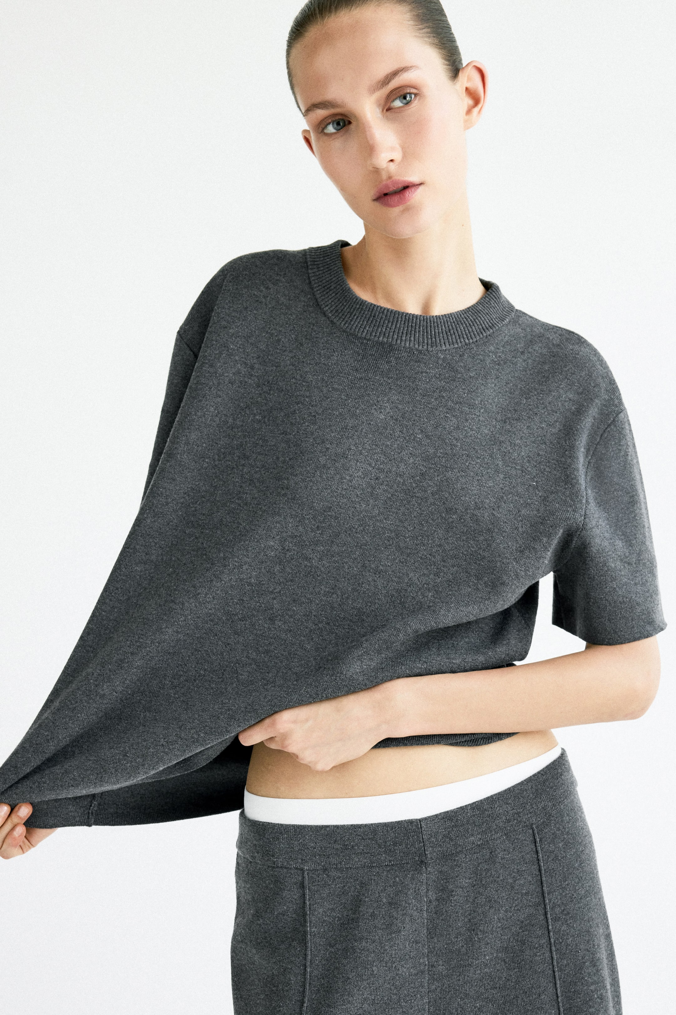 Ampliar la imagen: La modelo lleva puesta una camiseta extragrande de un tejido suave y fino en color gris oscuro, que presenta un escote redondo con ribete, hombros caídos y un dobladillo recto. La modelo sostiene el borde inferior de la camiseta con ambas manos, revelando su abdomen y la parte superior de un pantalón de punto a juego.