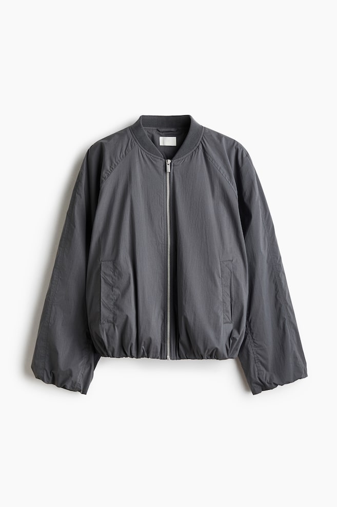 A dark grey bomber jacket is shown from the front, featuring a ribbed collar, a central silver zip closure, and wide raglan sleeves. It has vertical welt pockets with quilted stitching and an elasticated hem, creating a gathered, relaxed silhouette. - 1
