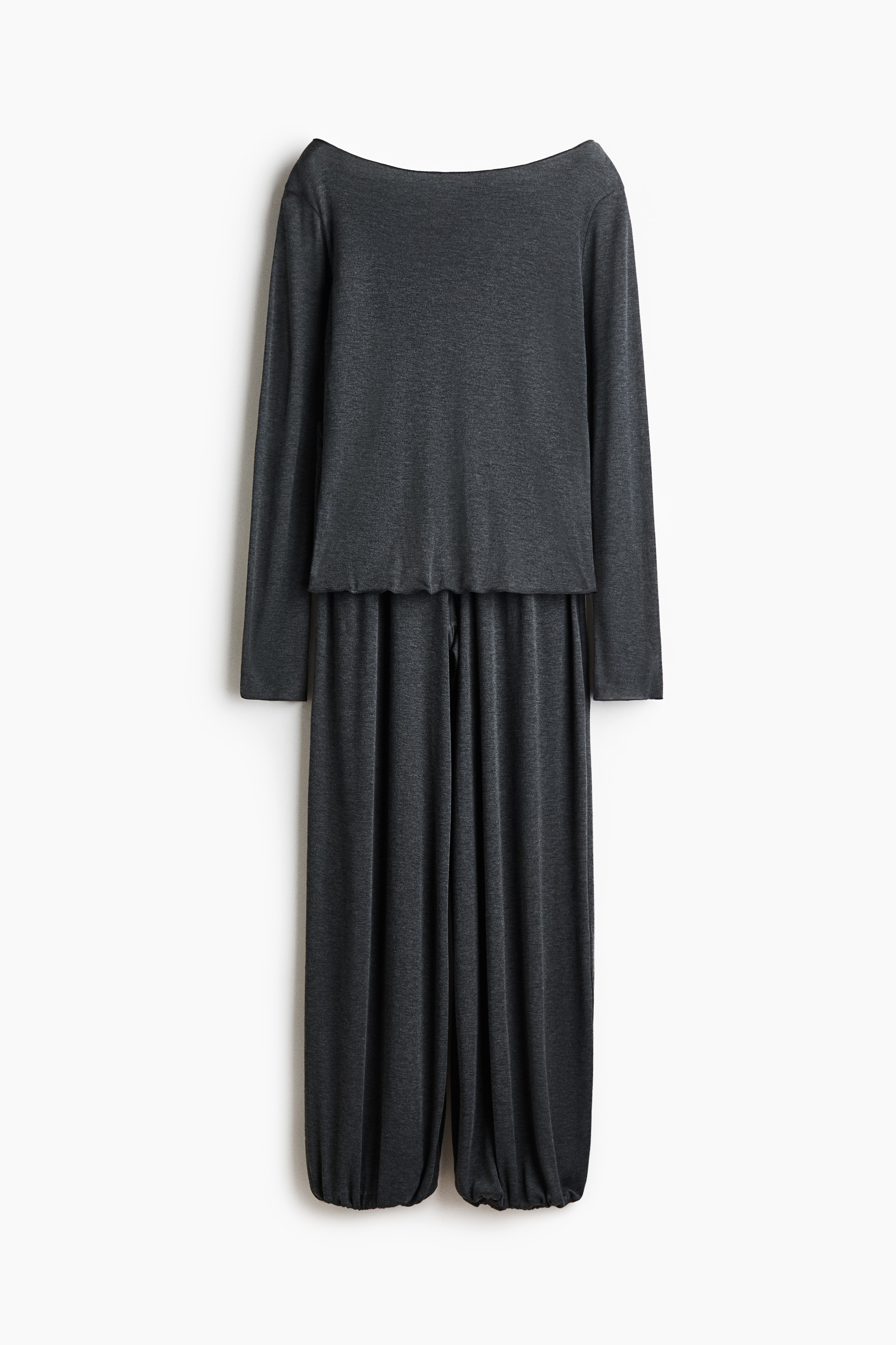 The model is wearing a dark grey jersey pyjama set, featuring a long-sleeved top with a boat neckline and matching loose-fit pants. The pants come with an elasticated waistband, a mock fly with three small decorative buttons, and balloon-style legs with elasticated hems.