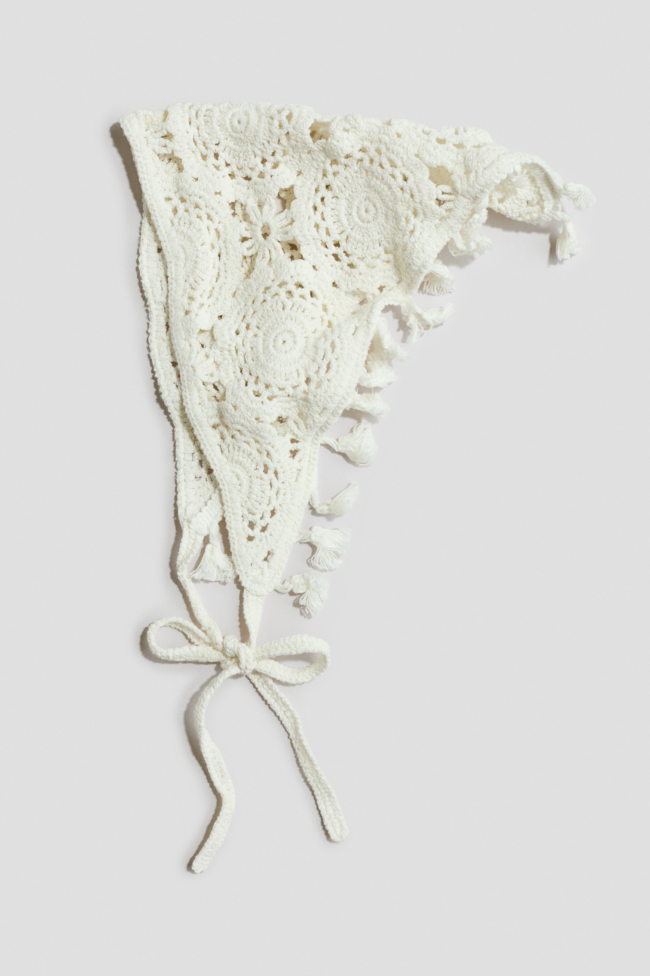 Voir une image plus grande: Un foulard blanc en crochet présente des motifs circulaires complexes et des ajourés, avec de petits glands ornant son bord festonné. Des liens en crochet s'étendent à partir d'un coin, noués en un nœud.