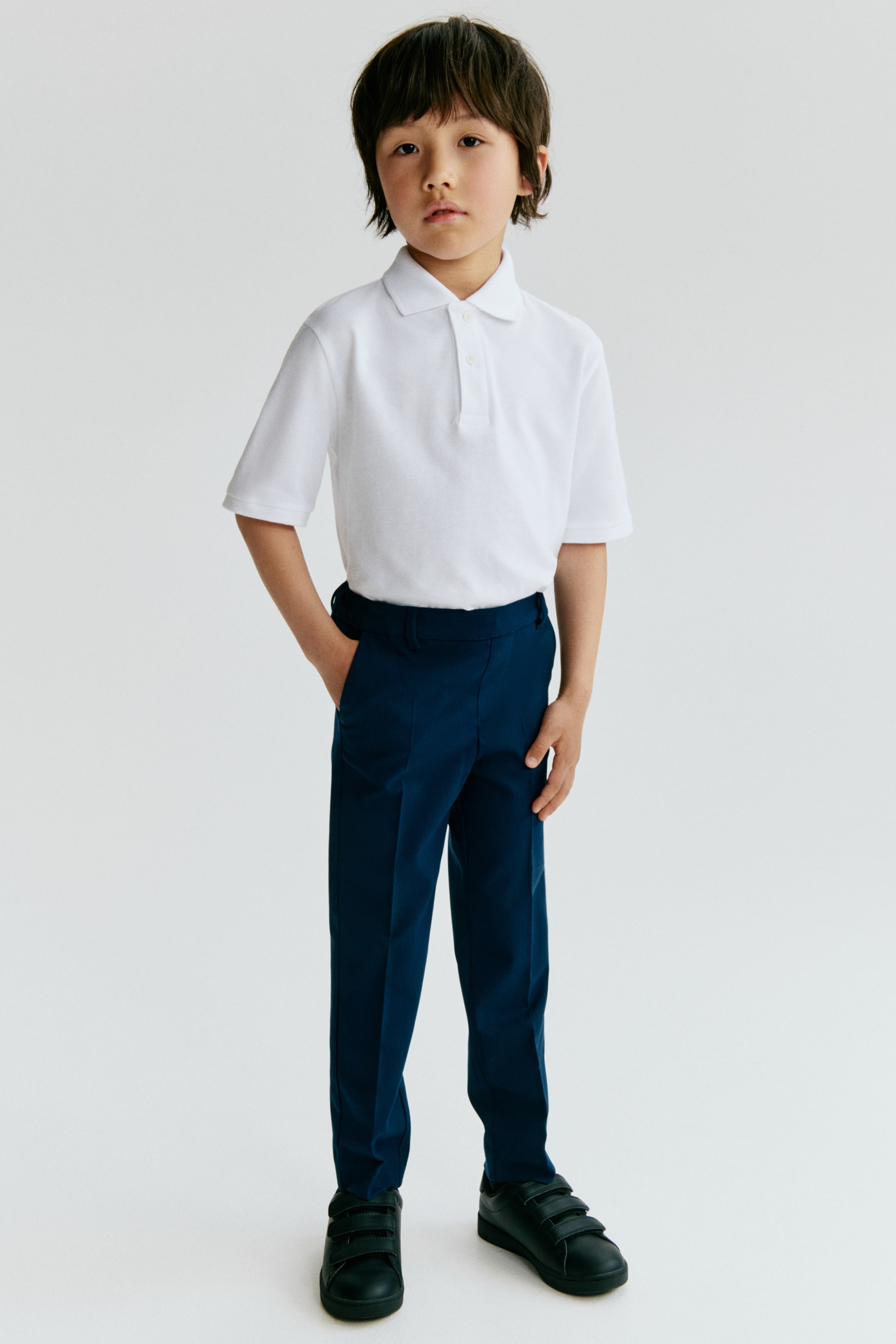 Ver imagen más grande: Una camiseta polo blanca de manga corta con cuello acanalado y una tapeta de dos botones se presenta desde el frente, y se lleva con unos pantalones azul marino de frente plano con pliegues a lo largo de las piernas y bolsillos laterales.