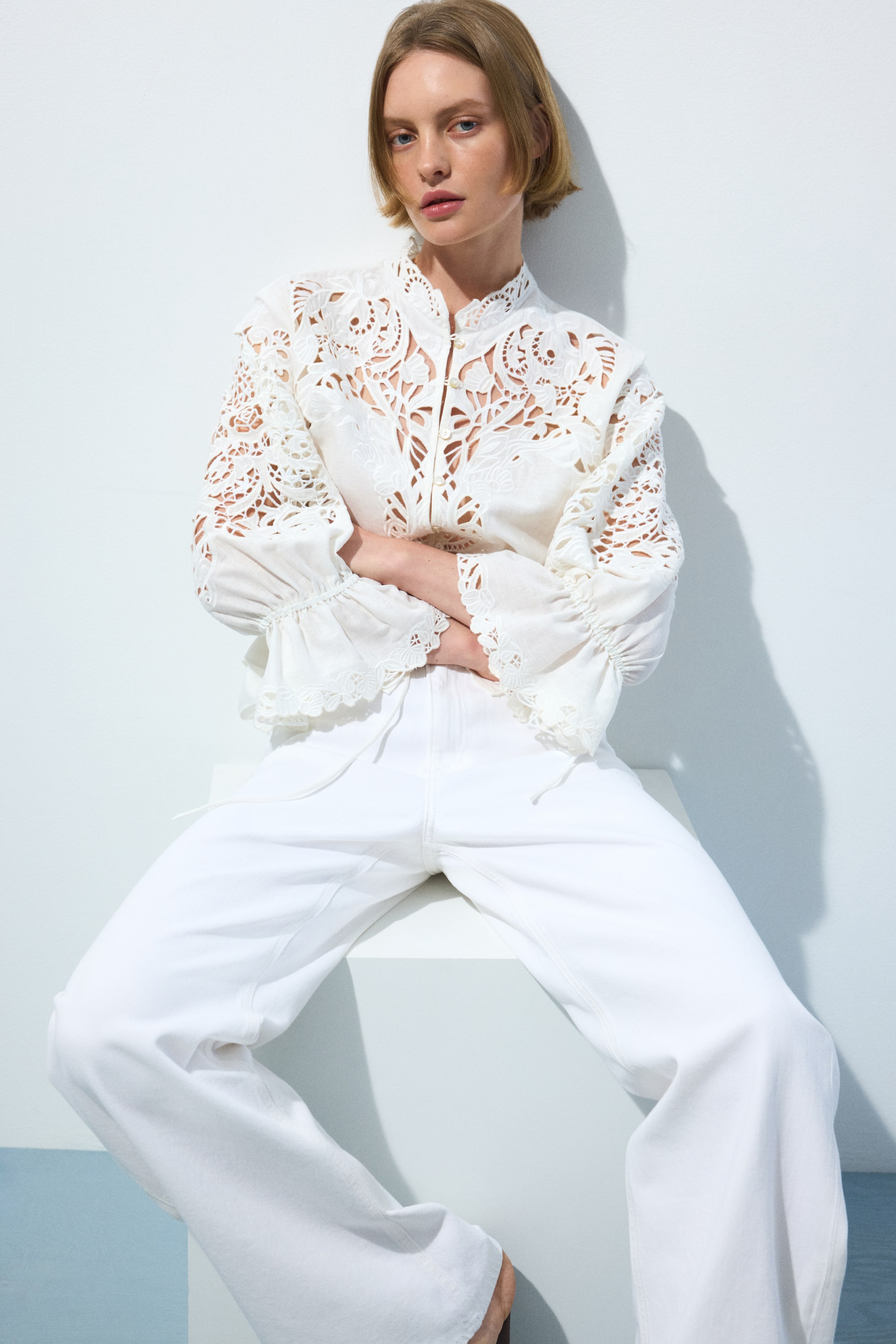 הצגת תמונה גדולה יותר: חולצה לבנה עם צווארון עומד ורקמת גזירה מורכבת על החזה, הכתפיים והשרוולים הארוכים, המסתיימים בחפתים מקופלים ומעוטרים. החולצה משולבת עם מכנסיים לבנים בגזרה גבוהה, המבליטים שרוך גלוי וגזרה רחבה ונינוחה.