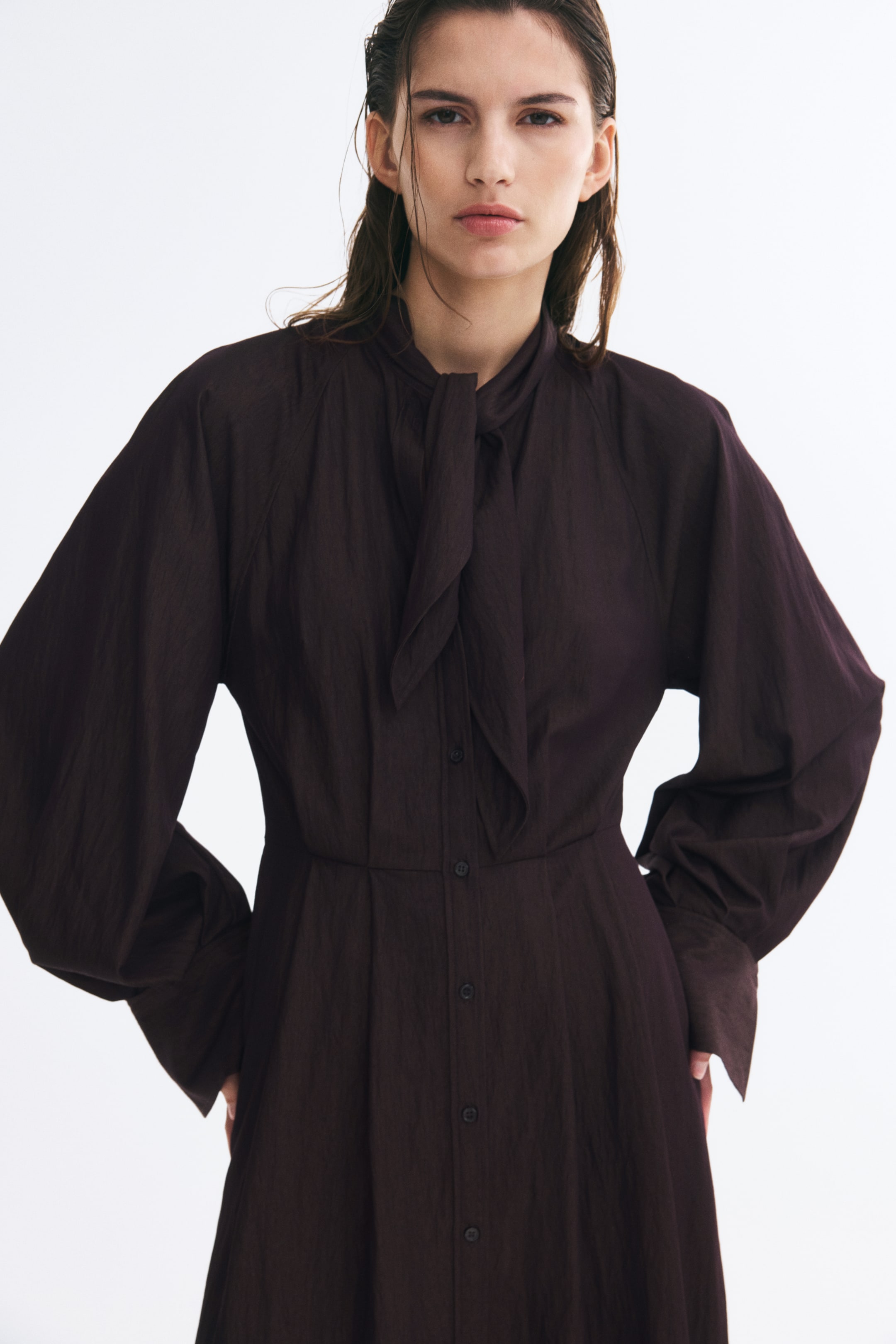 Ampliar la imagen: La modelo lleva puesto un vestido marrón oscuro de tejido texturizado. El vestido presenta un cuello tipo pañuelo anudado, cierre frontal con botones, costura marcada en la cintura y mangas largas abullonadas con puños anchos.