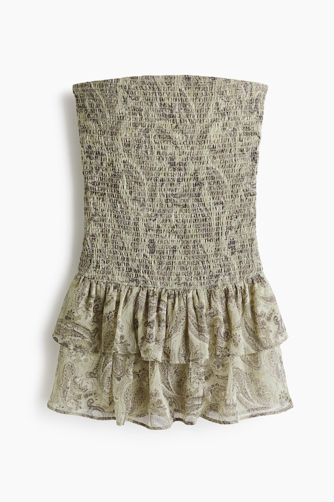 A short bandeau dress is facing front, featuring a smocked bodice. The dress is continuing with a gently flared skirt, displaying two flounced tiers, all in a light green fabric with a brown paisley pattern. - 1