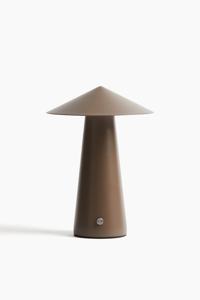 Současná dobíjecí stolní lampa stojí čelem dopředu, má matný, světle hnědý, kuželovitý podstavec, který se zužuje směrem nahoru a podpírá široký, plochý, kuželovitý stínidlo. Malé, kulaté, kovové tlačítko je umístěno na dolní přední části podstavce, nad tenkým černým kroužkem na spodním okraji. - 1