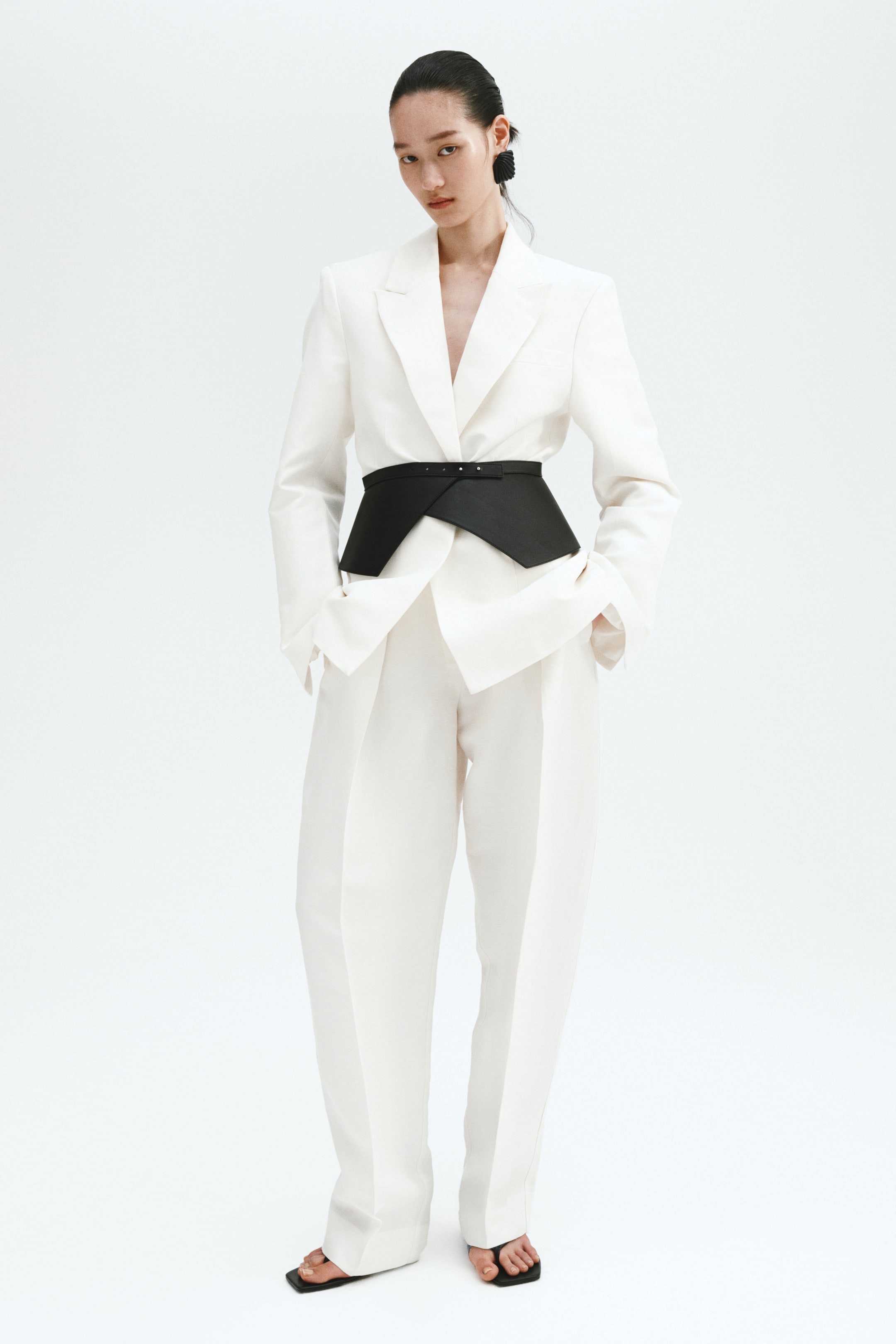 View larger image: A white tailored suit, comprising a blazer with wide notch lapels and structured shoulders, is paired with matching wide-leg trousers. A black asymmetrical corset belt adorned with small silver studs cinches the blazer at the waist.