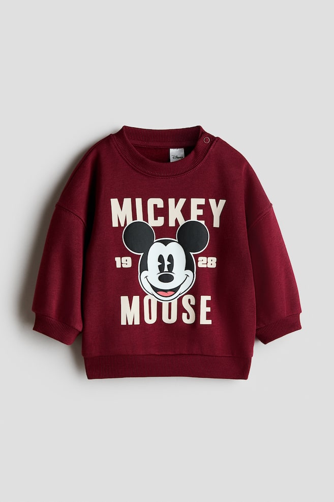 A maroon crew neck sweatshirt is lying flat, showcasing a large white graphic print on its front. The print displays "MICKEY" at the top, a smiling Mickey Mouse head with "19" and "28" on either side, and "MOUSE" at the bottom. The sweatshirt also features ribbed cuffs and hem, with two snap buttons visible on the left shoulder. - 1