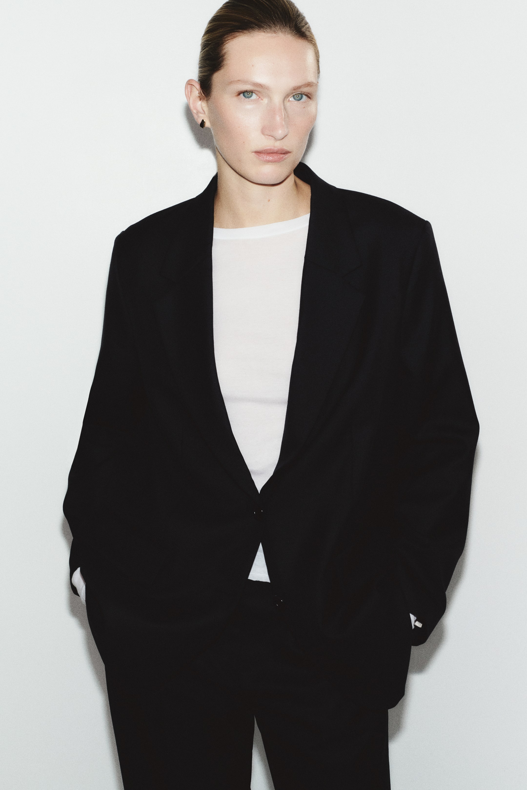 View larger image: A long black single-breasted blazer with notch lapels and a single-button front closure is styled on the model over a white T-shirt and matching black trousers. The model is standing with hands in the trouser pockets.