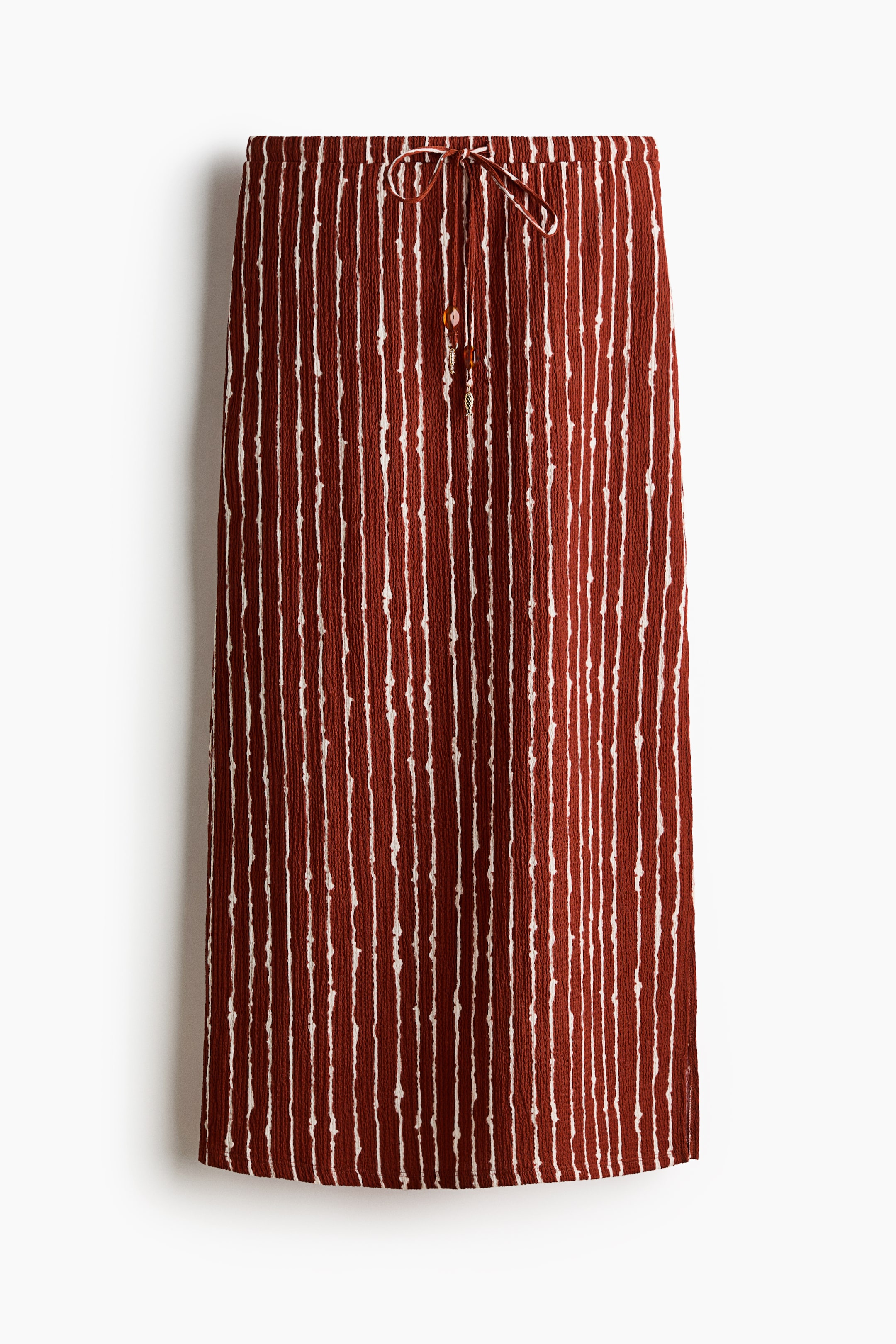 View larger image: A calf-length, fitted skirt is showcased in a crinkled dark red jersey with irregular vertical white stripes. It features a high, elasticated waist with a drawstring tied in a front bow, its ends adorned with a bead and a fish-shaped charm, and a straight-cut hem with a high slit on the right side.